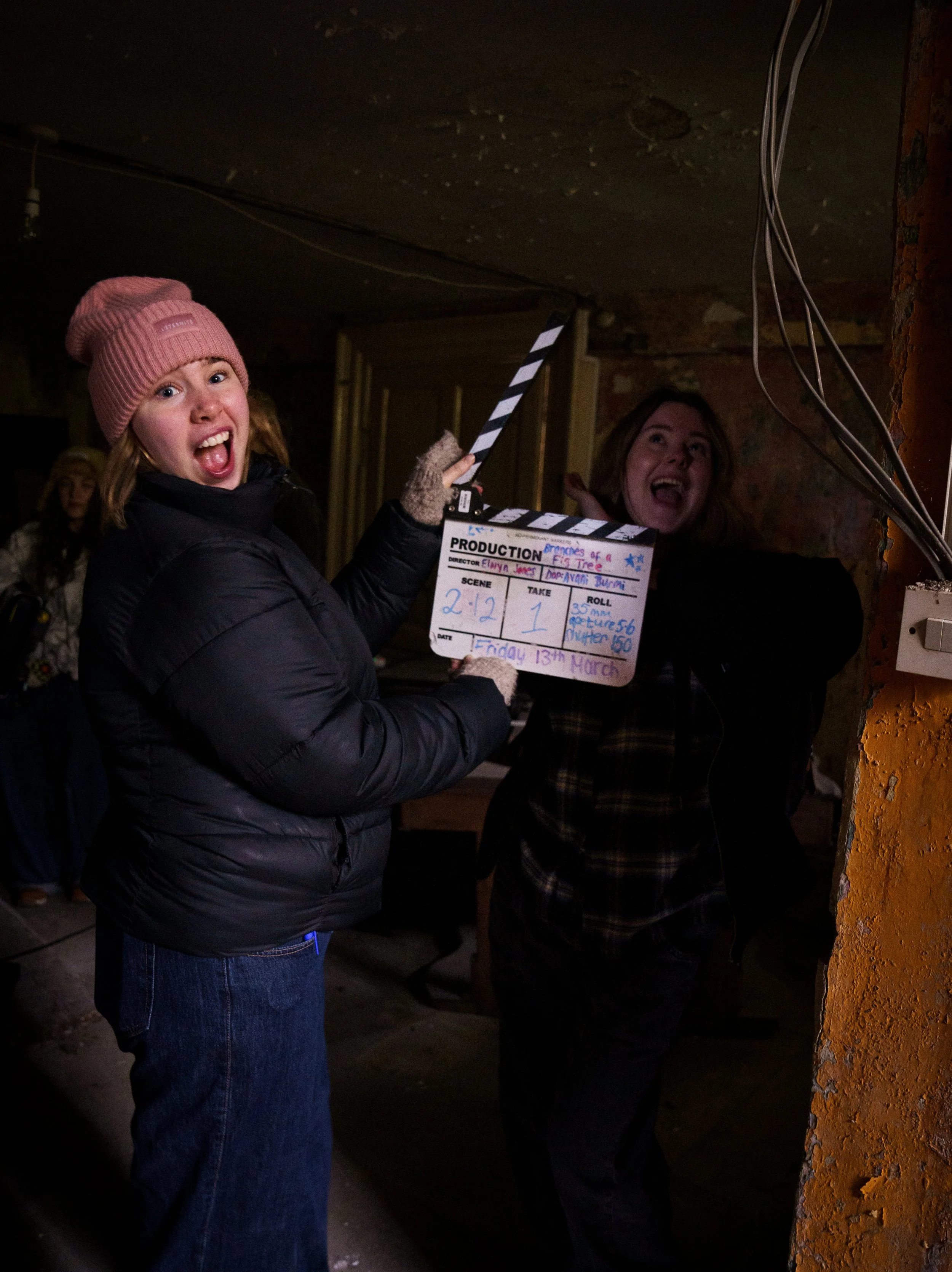 Two women on a film set, one holding a clapperboard, smiling and laughing in a dimly lit, rustic room.