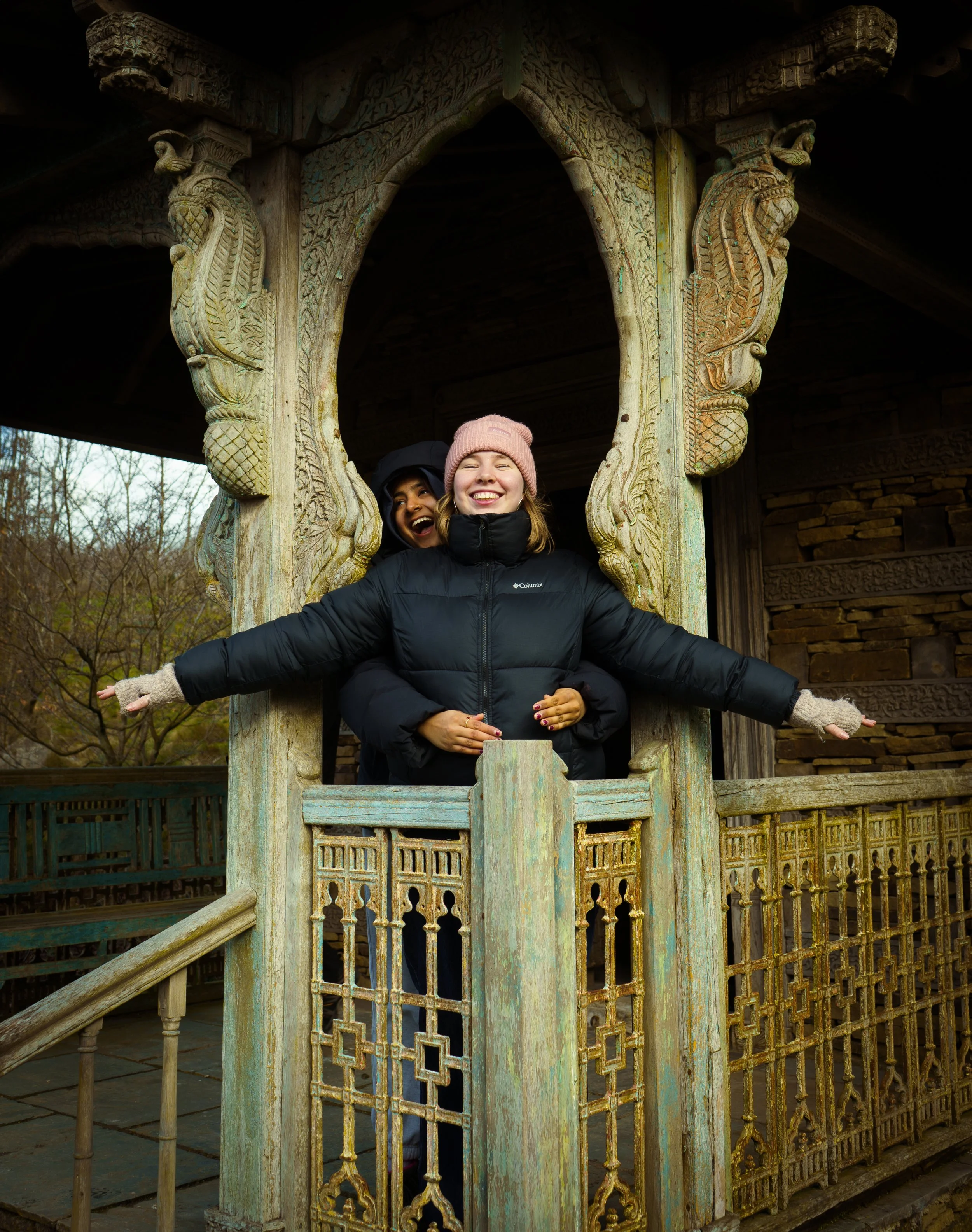 Two women smiling and posing in an ornate wooden pavilion outdoors. One is in front wearing a black jacket and pink knit hat, and the other is behind her, partially hidden, wearing a dark coat and hood.
