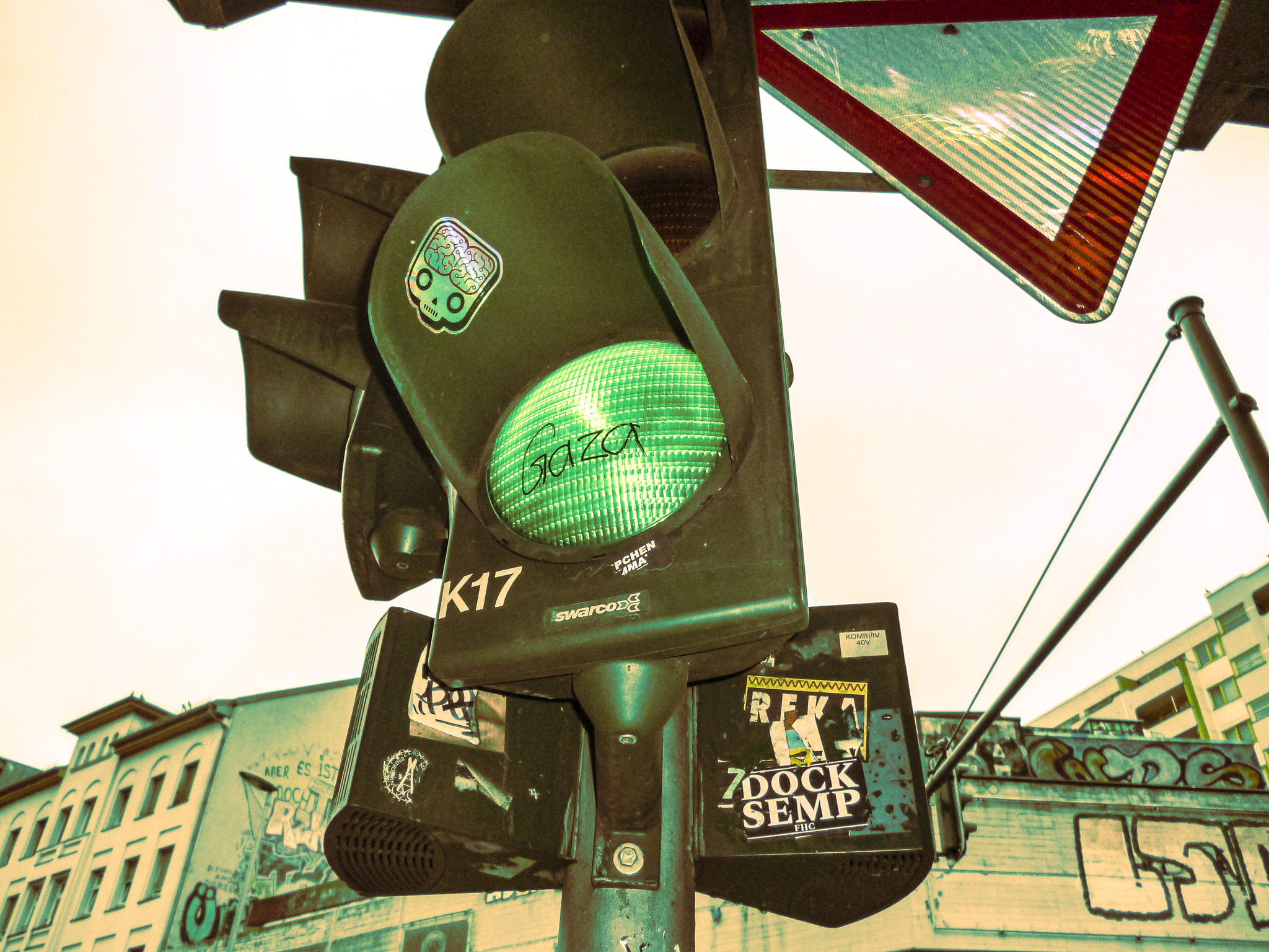 A traffic light with the green light on, displaying graffiti and stickers, located in an urban area with buildings covered in graffiti in the background.