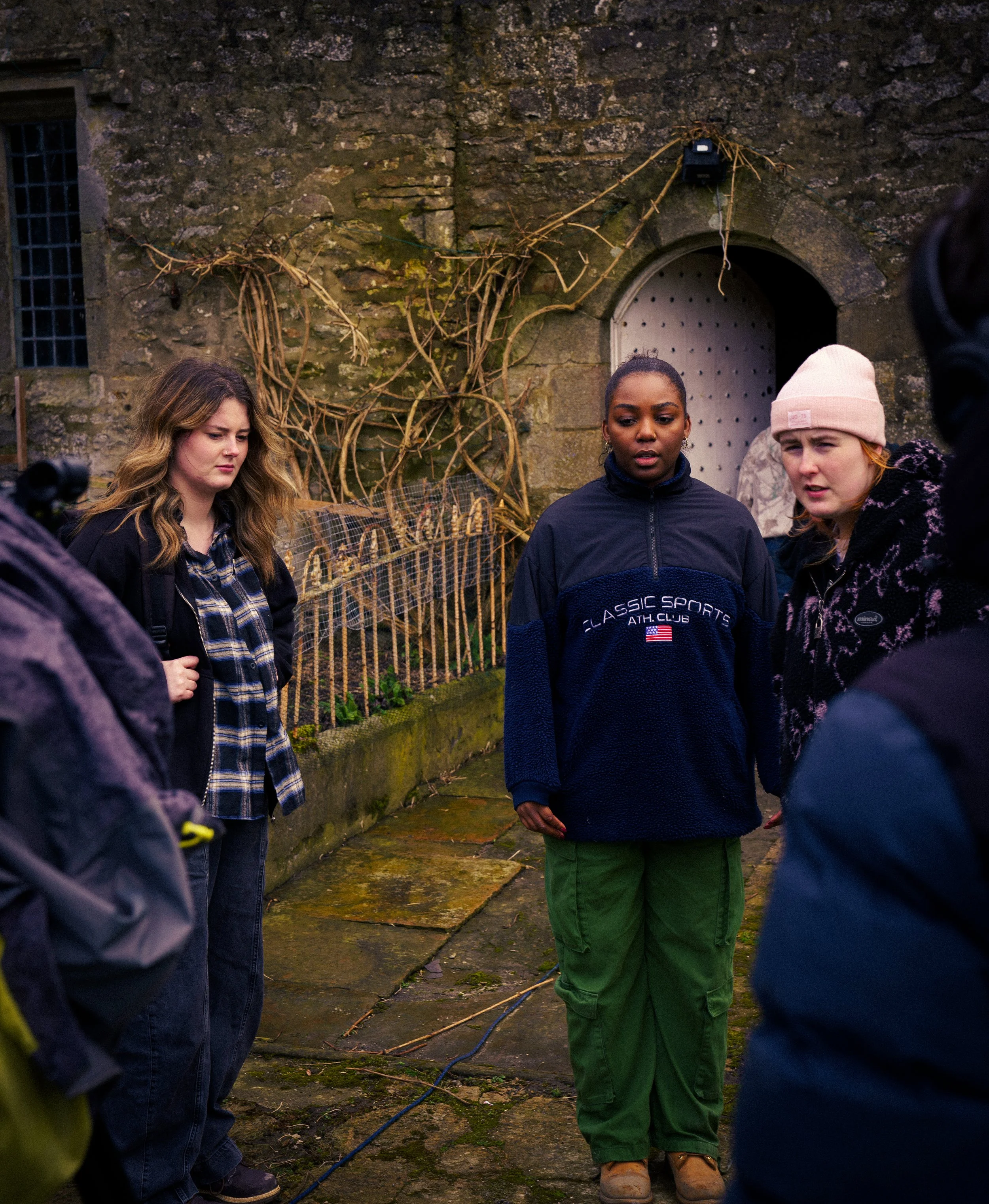 Group of young people standing outdoors on a mossy stone path, with a stone wall and tied vines in the background, engaged in a serious conversation.