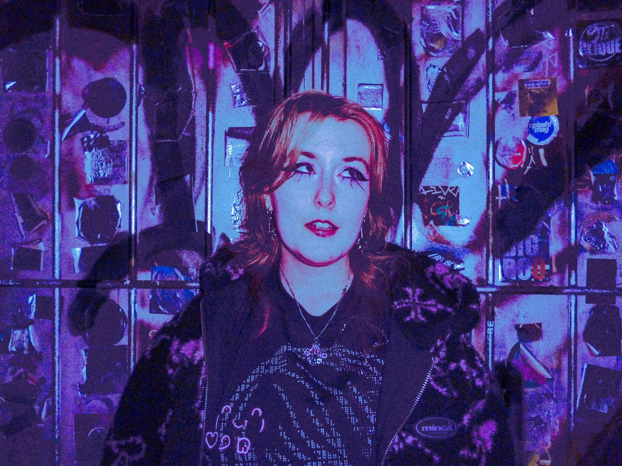 A woman with red hair, makeup, and wearing dark clothing with a necklace, standing in front of a wall covered with posters and stickers, illuminated by purple and blue lights.