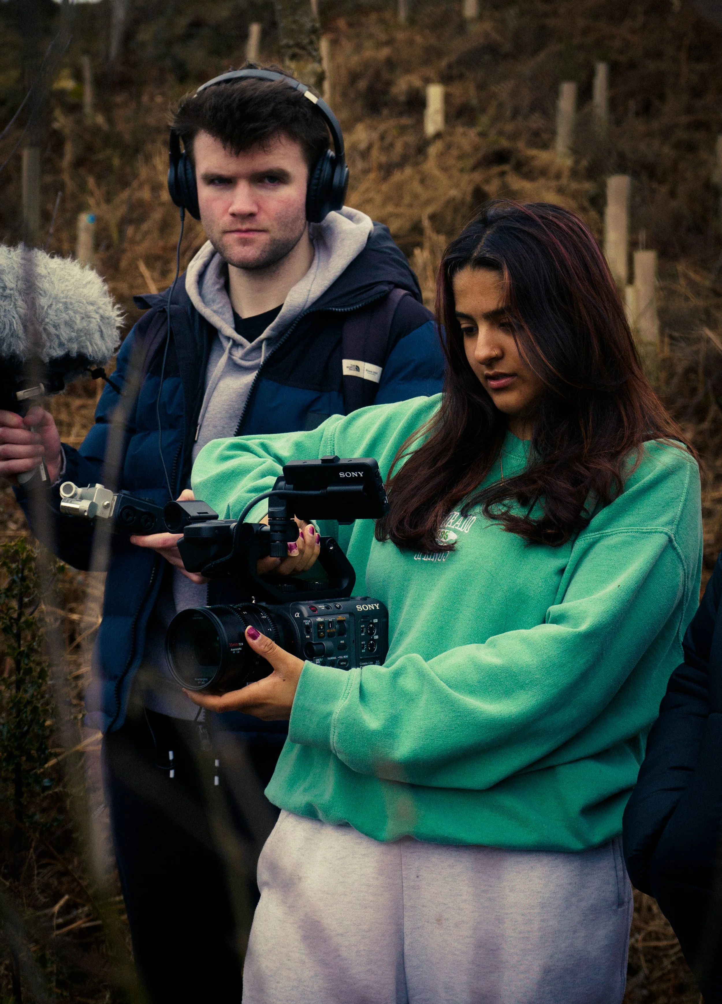 A woman operating a professional camera outdoors on a cloudy day, with a man with headphones holding a boom microphone nearby.