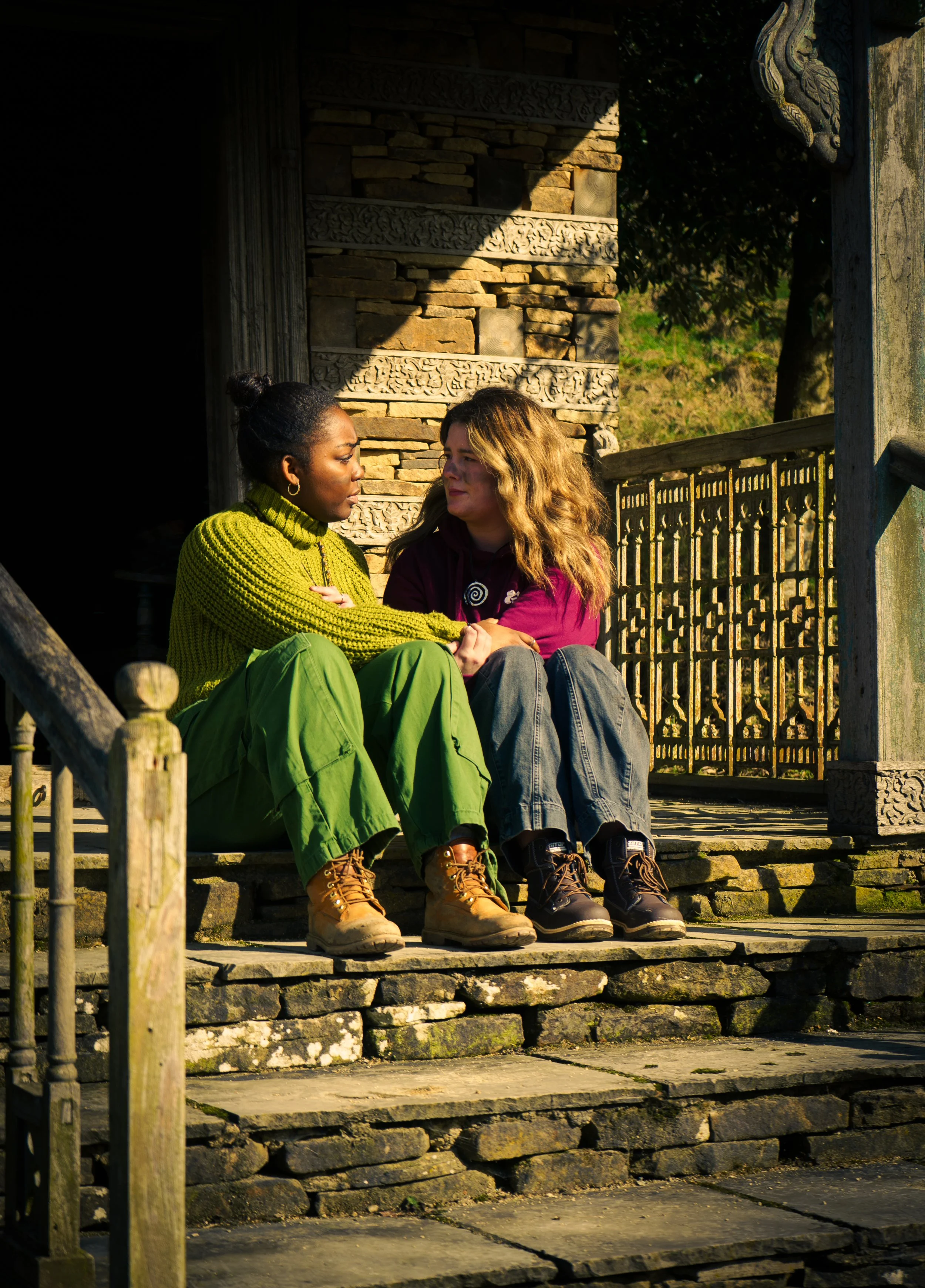 Two women sitting on stone steps outdoors, facing each other, engaged in a serious conversation. One woman has dark skin, is wearing a yellow-green sweater and green pants, with her hair in a bun. The other woman has light skin, is wearing a purple jacket and dark jeans, with blonde wavy hair.