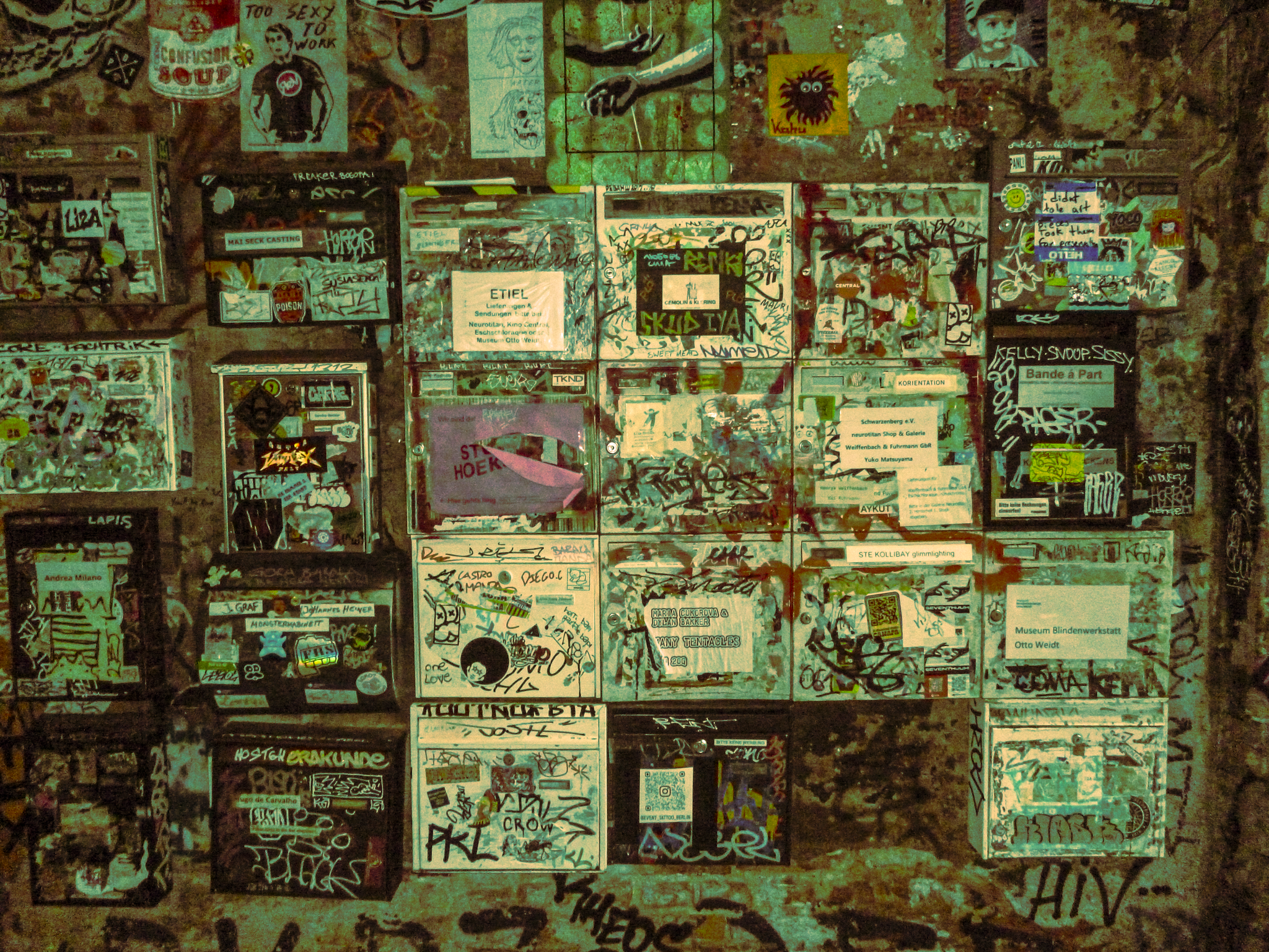 A wall covered with numerous graffiti, stickers, and posters, some with drawings or text. The wall has a rough, textured surface with various layers of graffiti overlapping each other.