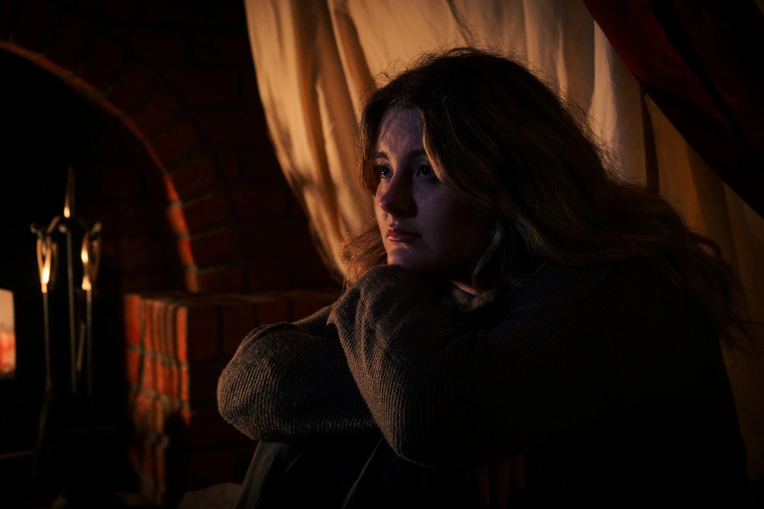 A woman with long brown hair sitting by a fireplace, resting her chin on her knees, with dim warm lighting and a brick wall background.