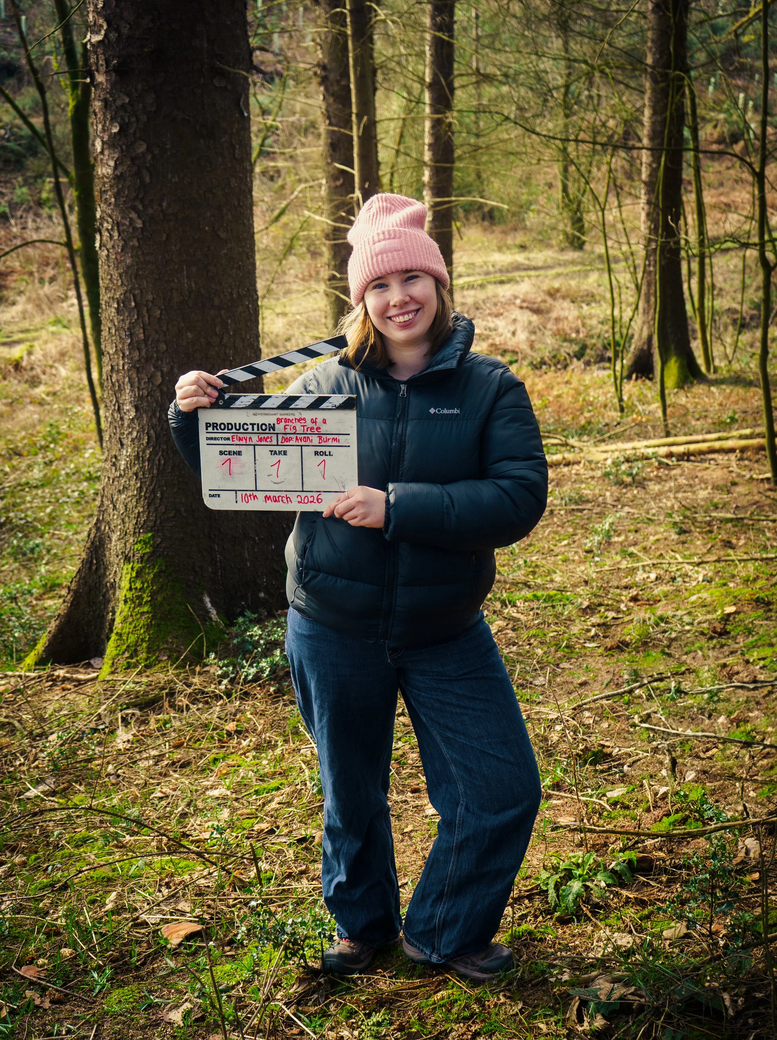 A woman in a pink knit hat and black puffer jacket standing in a forest, holding a clapperboard with the scene labeled "Branches of a Fig Tree," dated March 10, 2026, smiling at the camera.