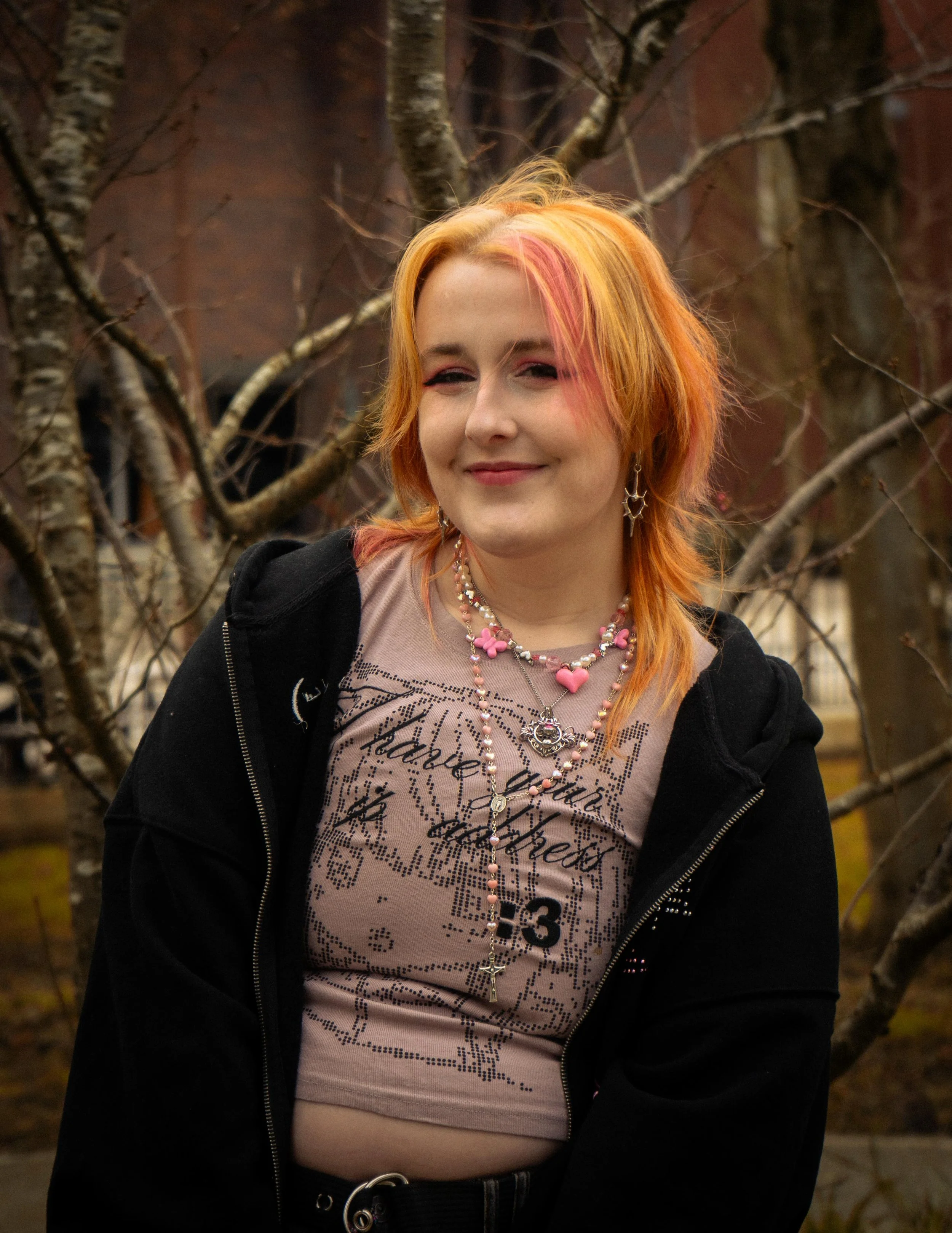 A young woman with shoulder-length orange hair with pink highlights, smiling outdoors in front of bare trees with a brick building in the background.
