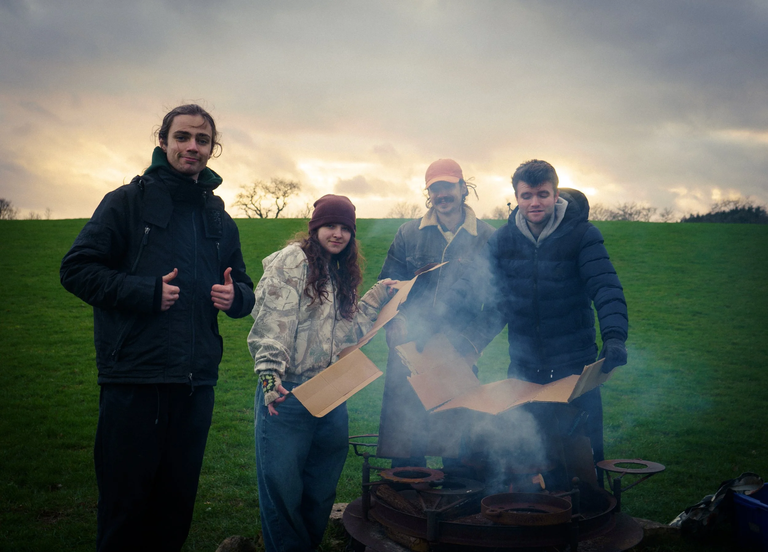 Four young adults standing outside around a campfire with smoke, on a grassy field under a cloudy sky at sunset.