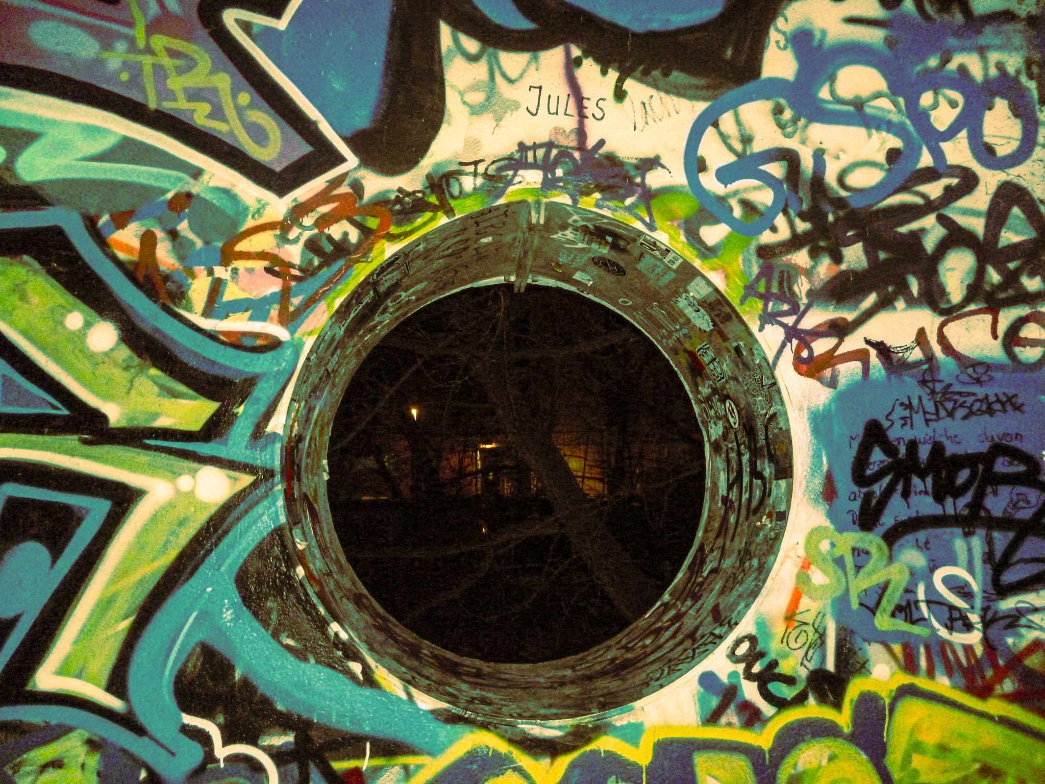 A graffiti-covered wall with a hole in the center that reveals a dark underground tunnel or space. The wall is decorated with colorful spray paint tags and designs.