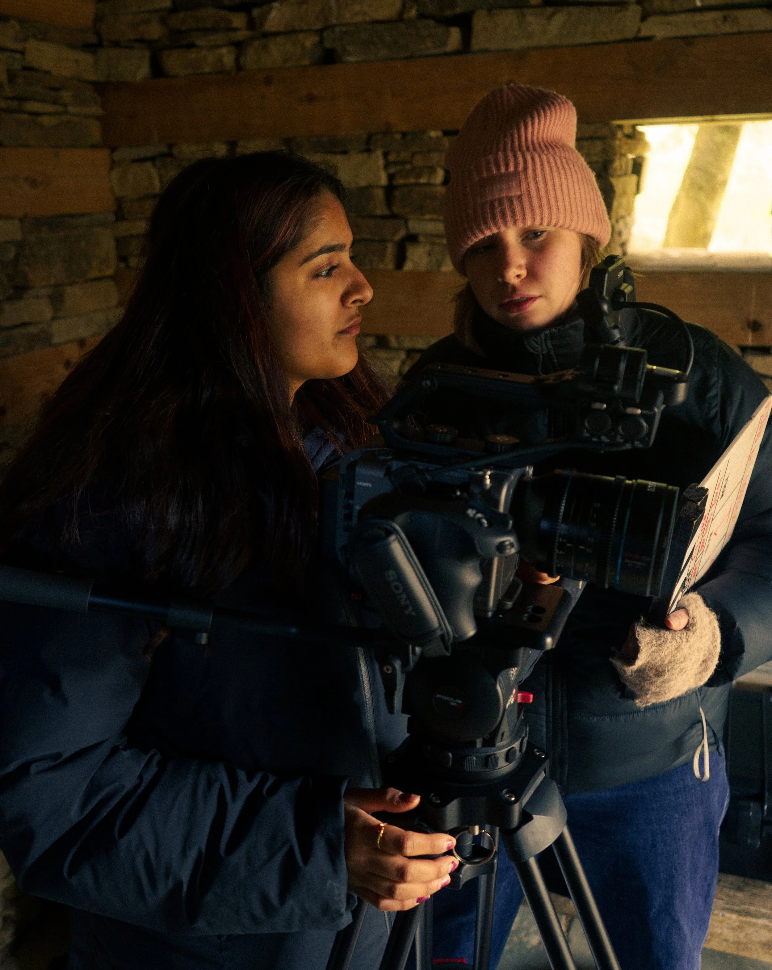 Two women on a film set, one with long dark hair and the other with a pink knit cap, inspecting camera and film equipment in a rustic setting.