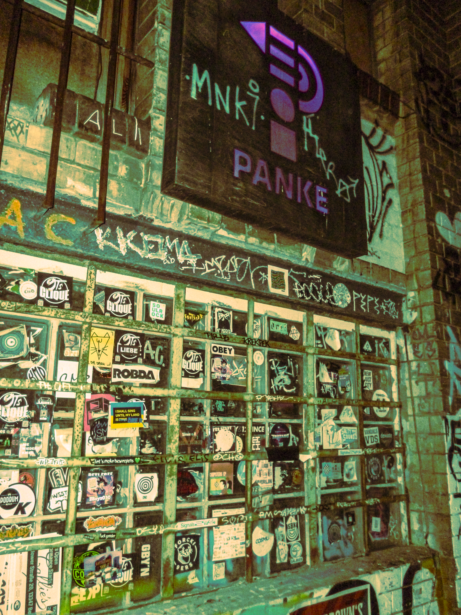 A wall with numerous stickers and graffiti, including a neon sign that says 'Panke' and other unreadable graffiti and stickers.