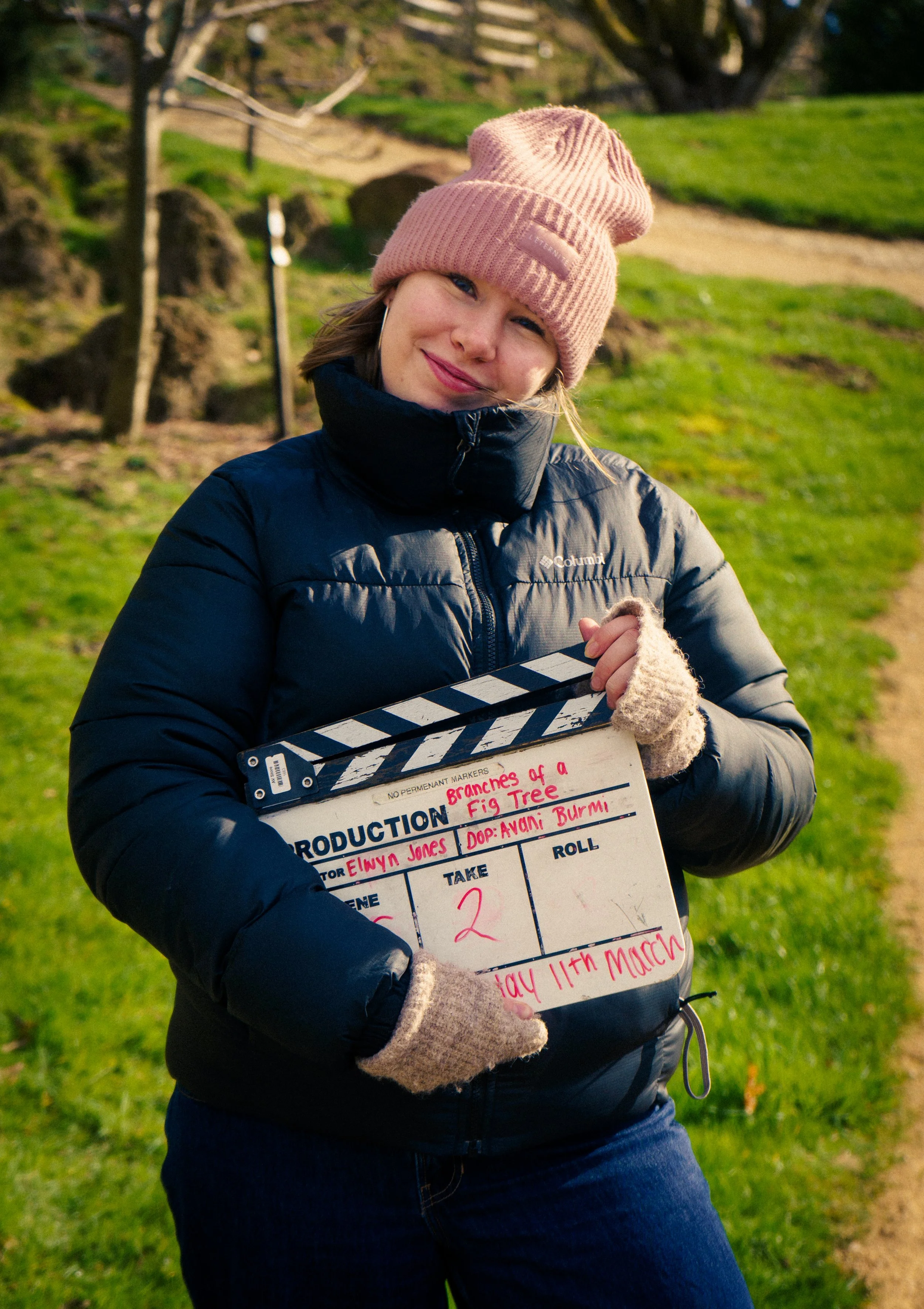 Woman in a pink beanie and navy jacket holding a film slate in a park with green grass and trees.
