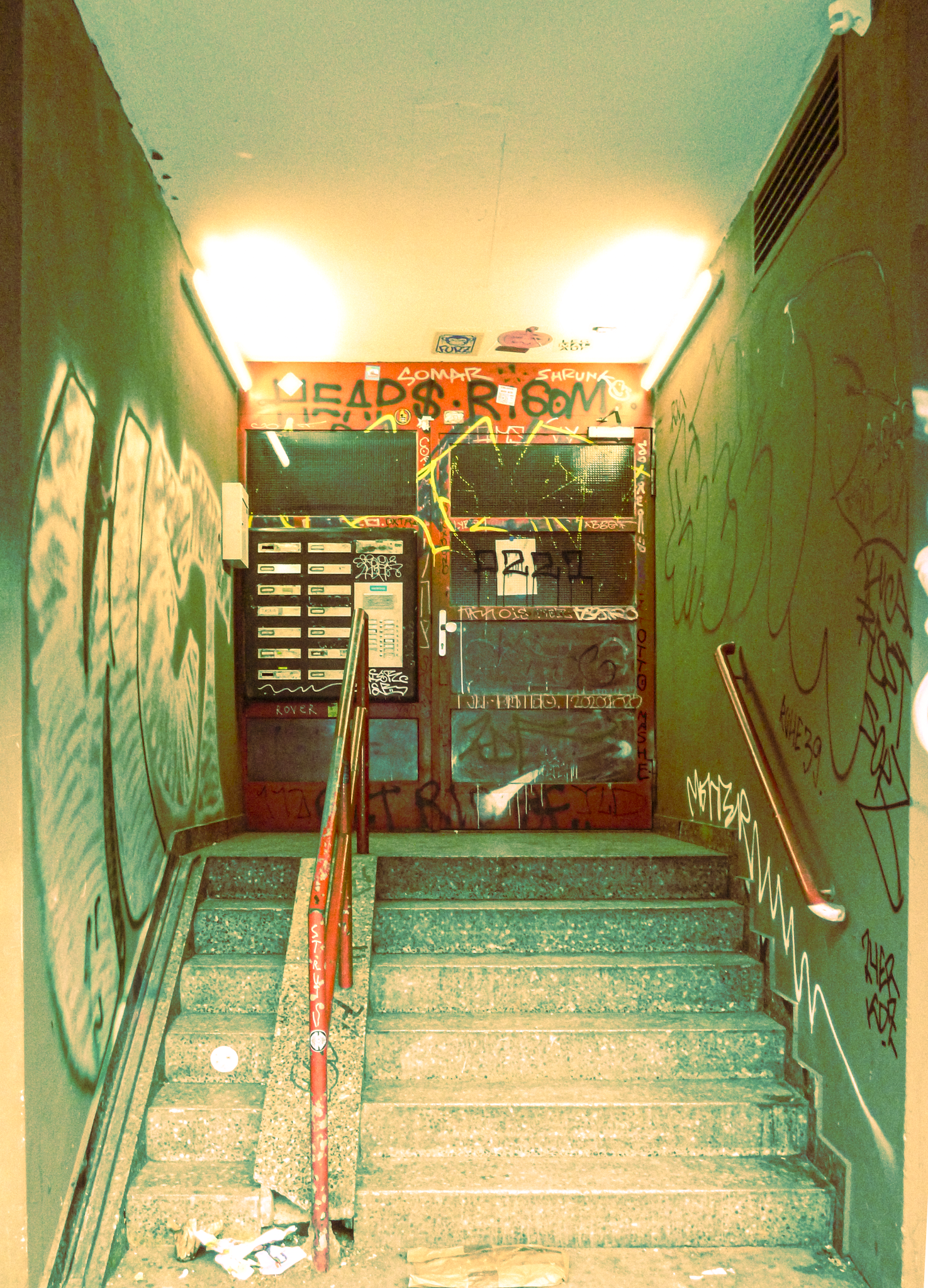 Graffiti-covered stairs leading to an entrance with a metallic gate, graffiti on the walls, and a handrail on the right side.