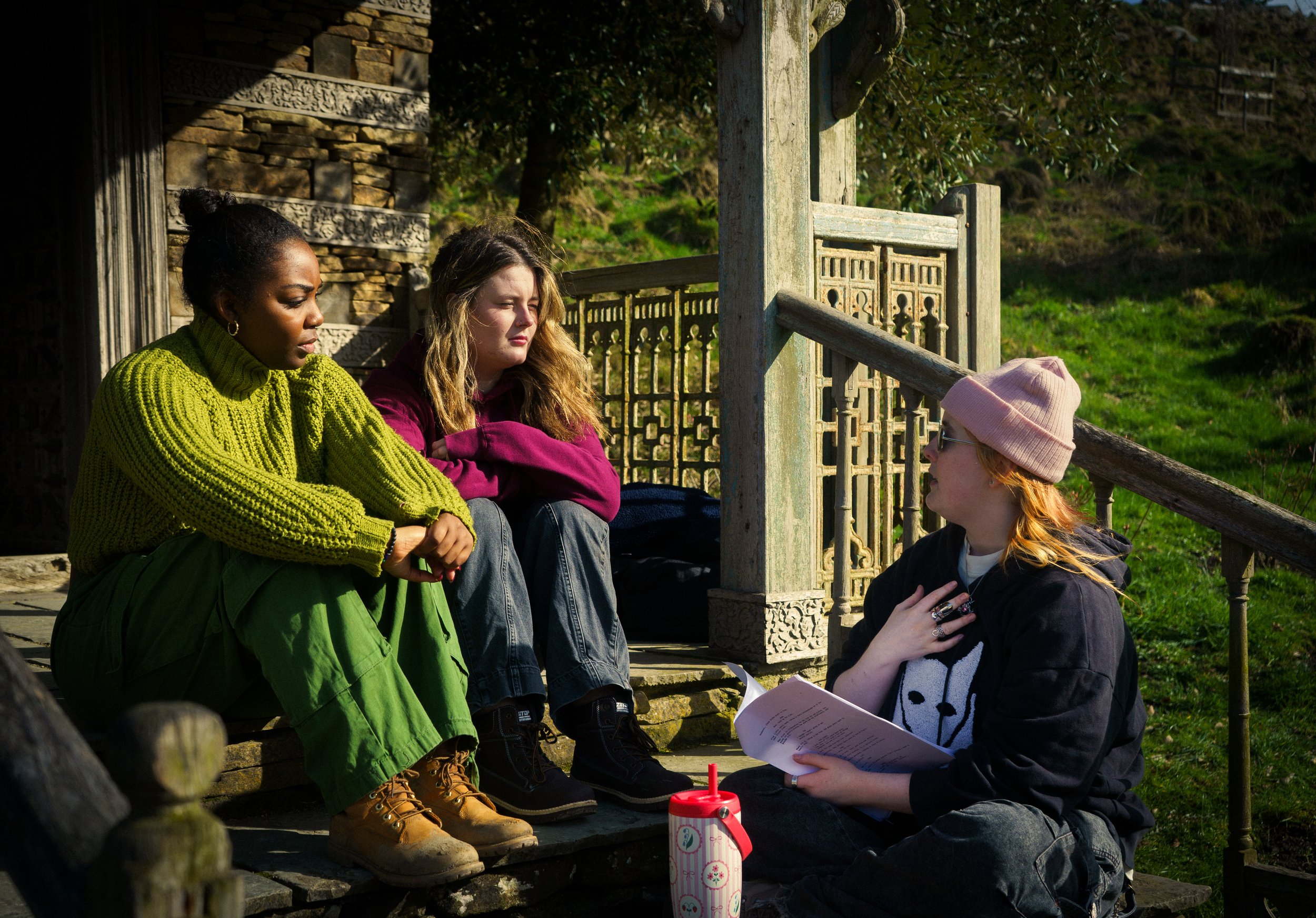 Three people sitting outdoors on a stone step having a conversation. One woman in a bright yellow sweater and green pants, one woman in a magenta jacket, and one woman with a pink beanie and black hoodie holding papers.