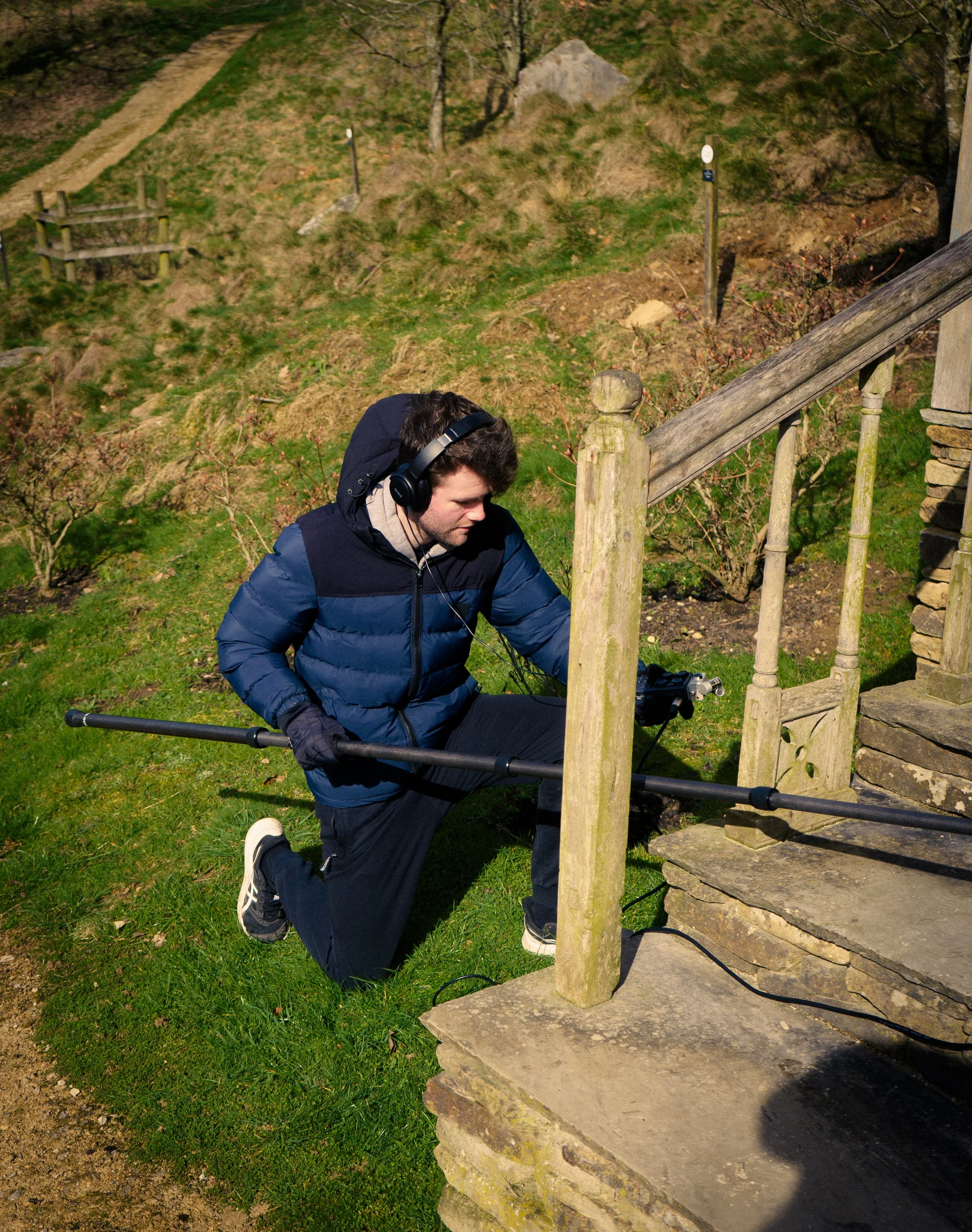 A man wearing a blue jacket, headphones, and gloves kneeling on grass, holding a long pole, and working on a wooden staircase outdoors in a grassy and hilly area.
