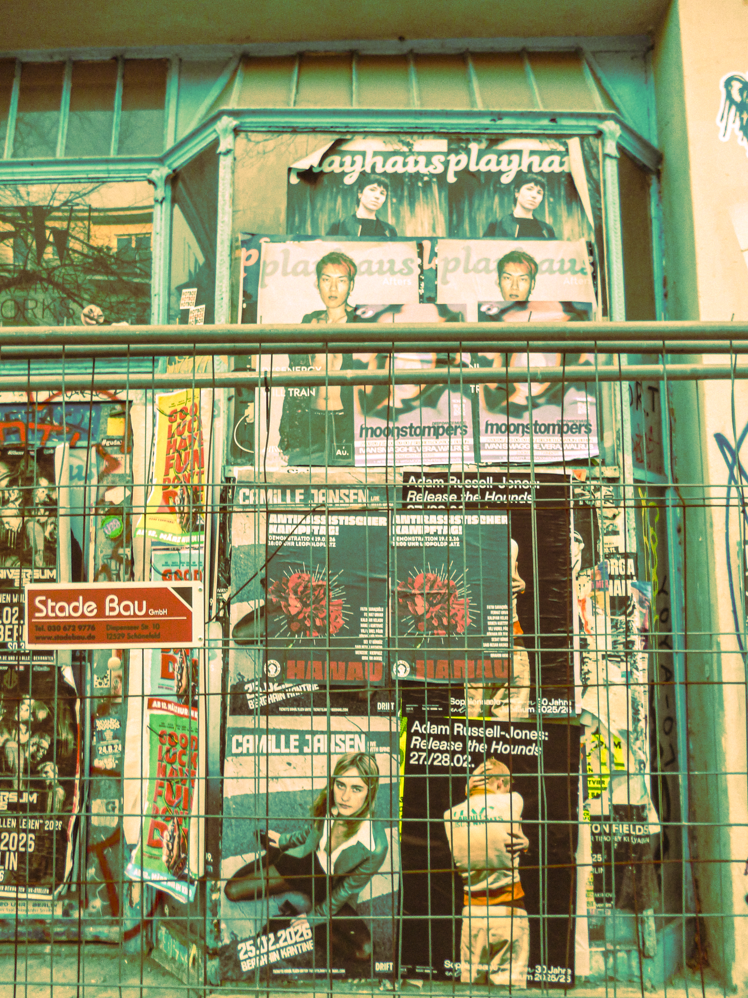 A wall covered with various posters advertising concerts and events, partially behind a metal fence.