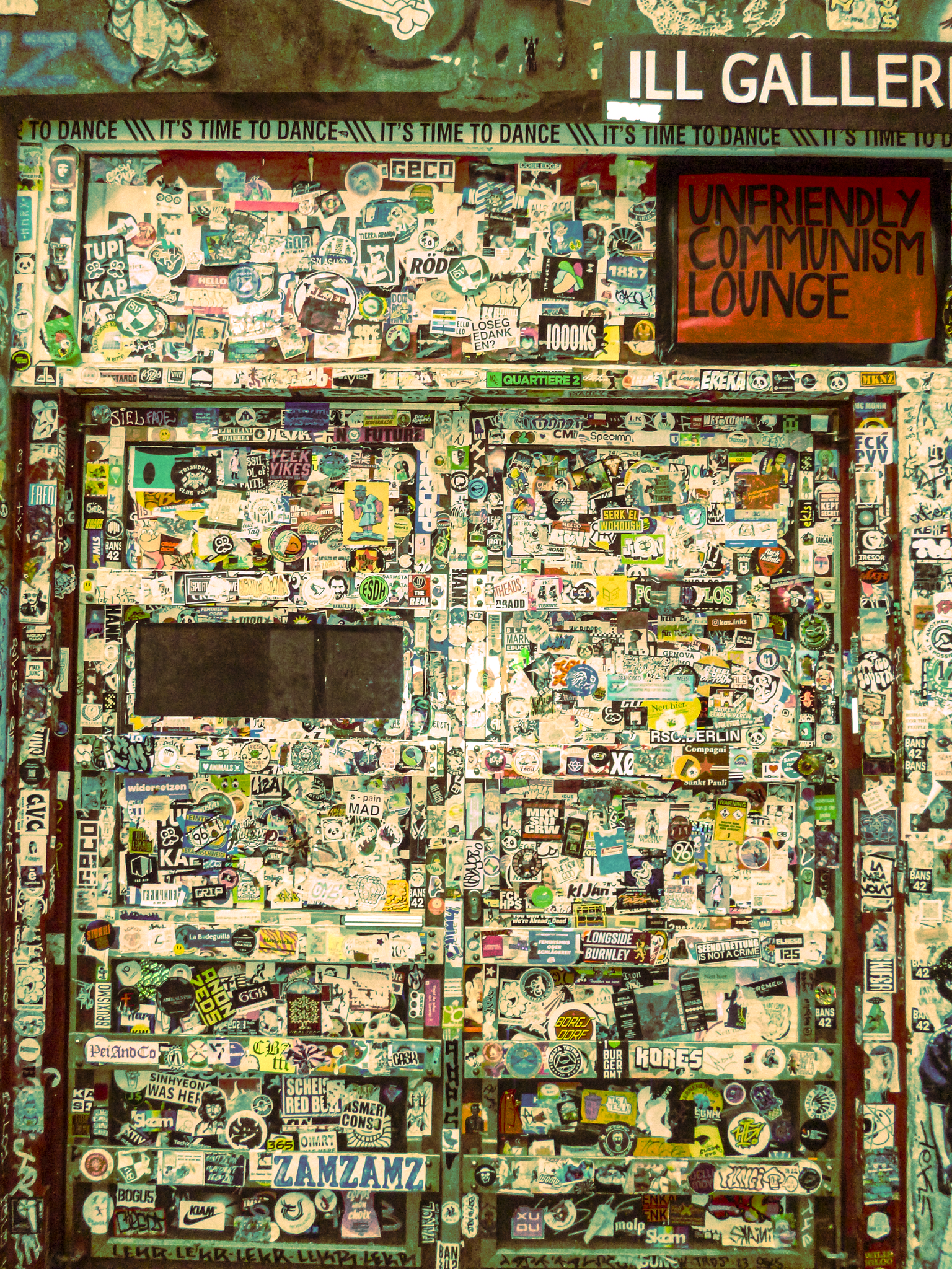 Colorful door covered in stickers and graffiti, with signs and a neon sign reading "Unfriendly Communism Lounge."