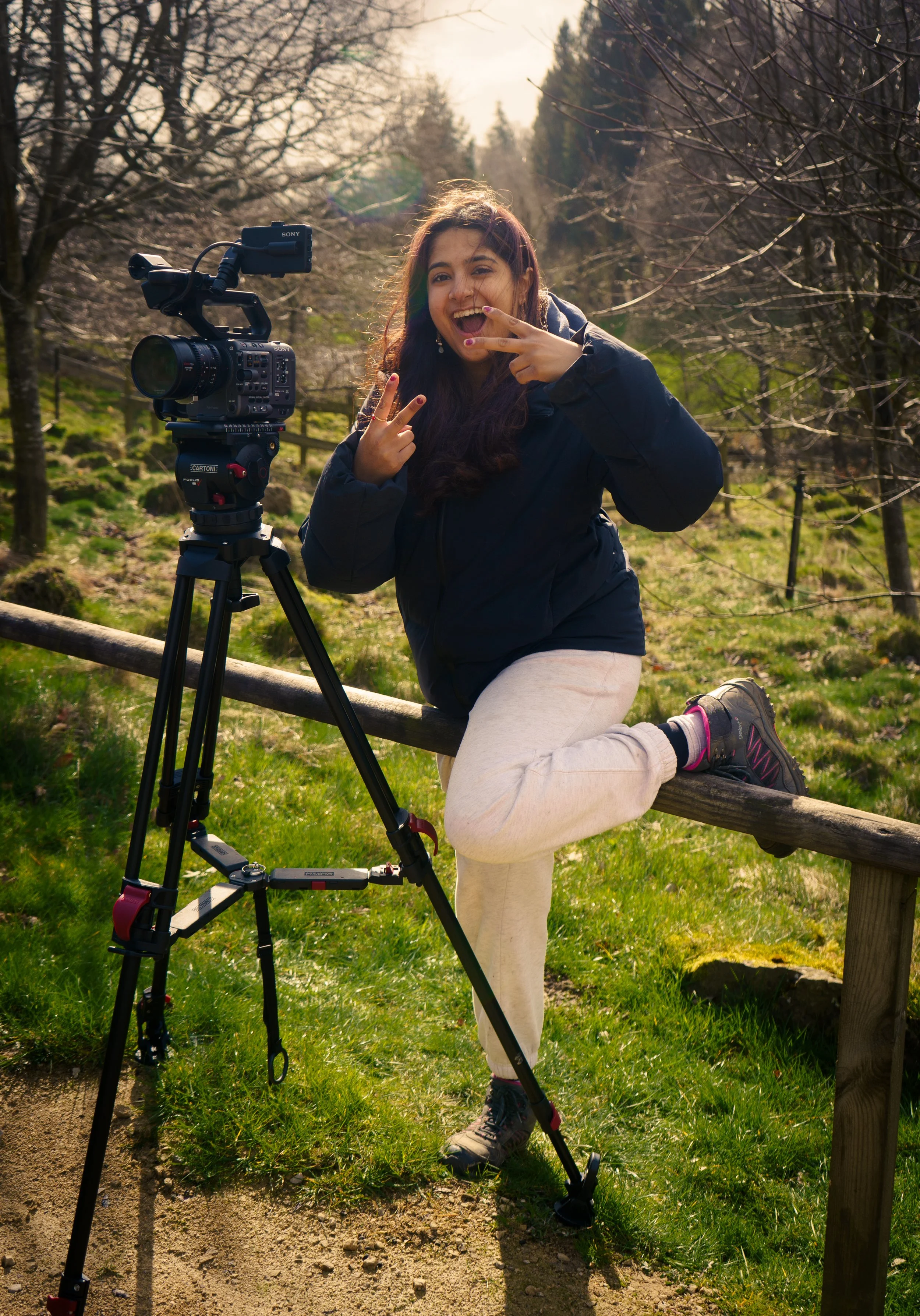 Young woman with long hair, wearing a dark jacket and beige pants, making a peace sign with her hands, kneeling on a wooden fence next to a camera on a tripod in a grassy outdoor area with leafless trees and sunlight.