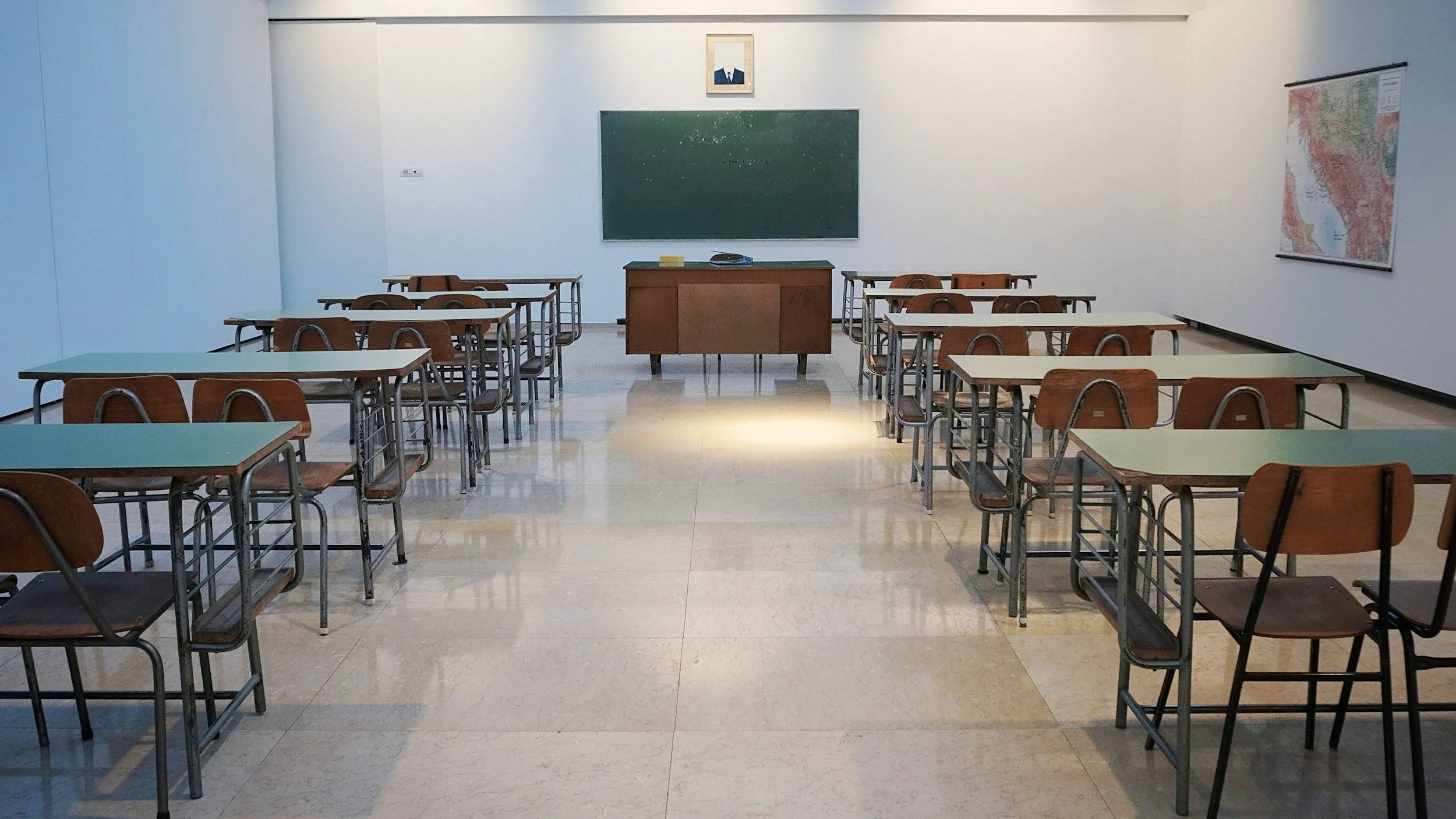 A classroom with rows of desks and chairs, a blackboard at the front, a framed photo above the blackboard, and a map of Greece on the wall to the right.
