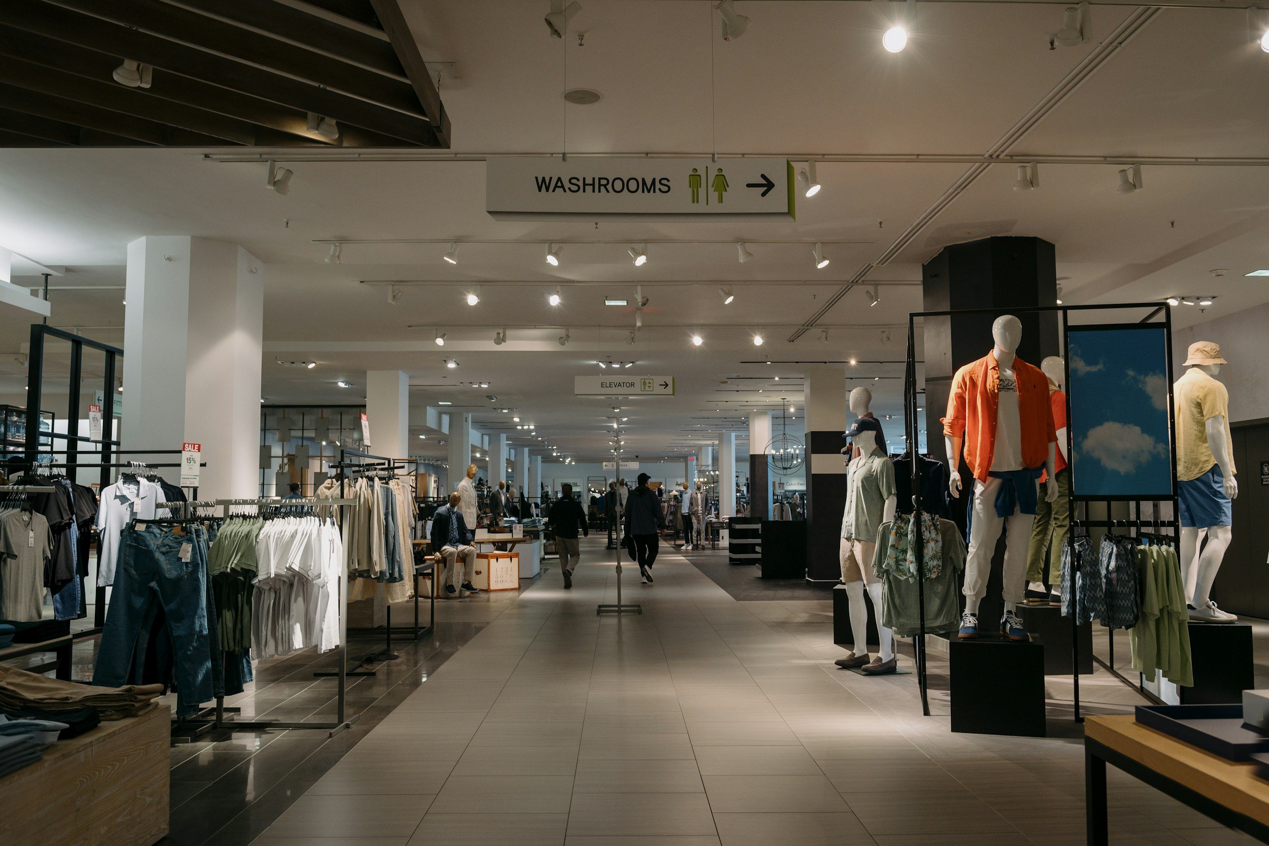 Interior of a clothing store with mannequins dressed in casual clothes, racks of clothes, and signs indicating washrooms and an elevator.
