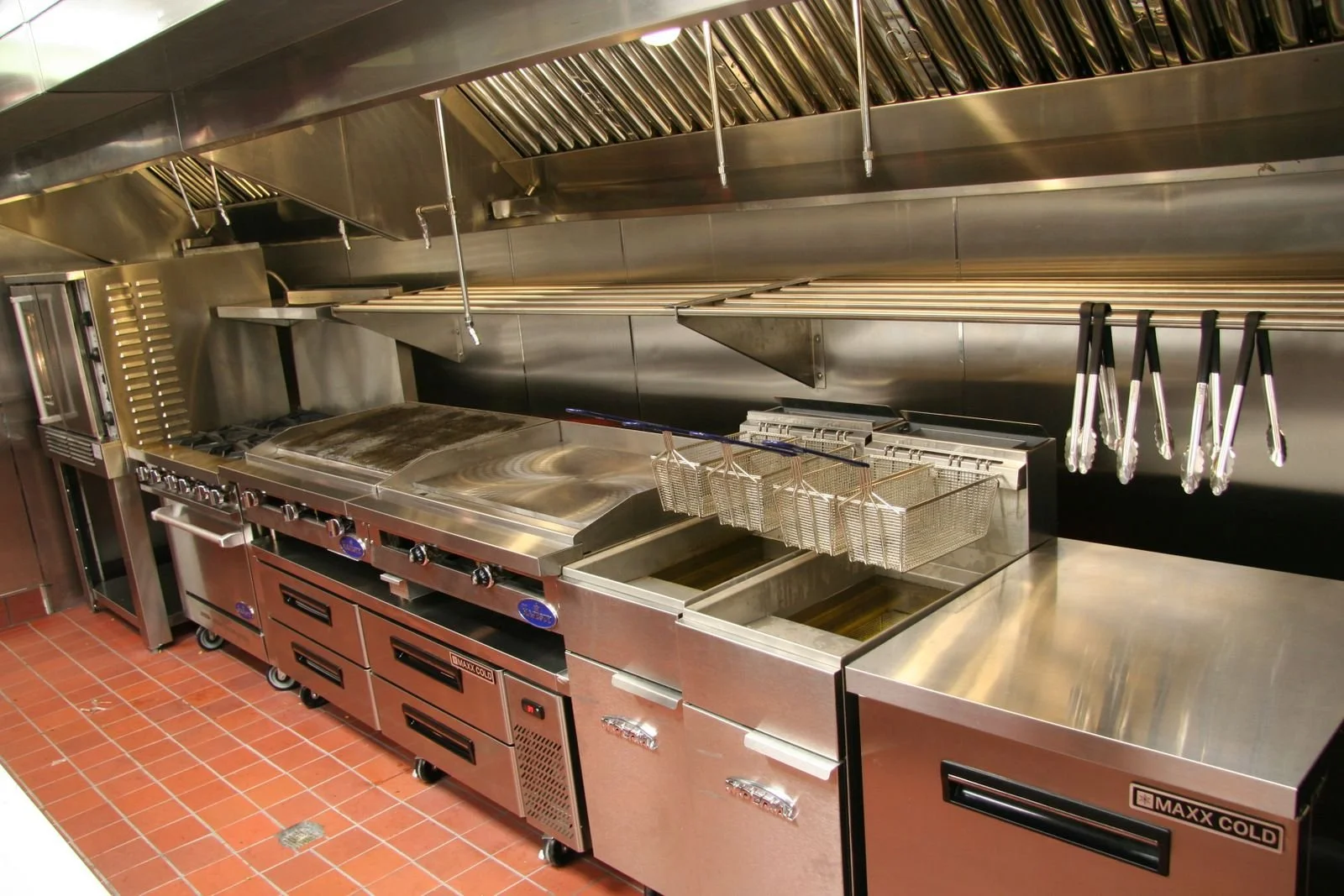 Commercial kitchen with stainless steel appliances including a flat-top griddle, fryers, and storage drawers. There are hanging kitchen knives and utensils on the right side and a ventilation hood above. The floor is made of red tiles.