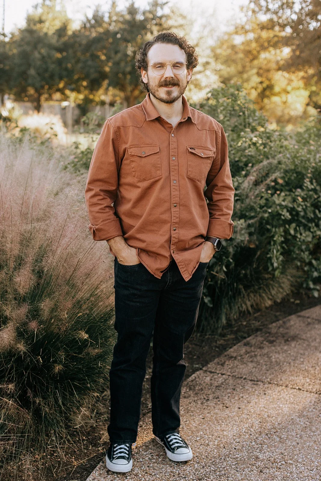 A man standing outdoors on a gravel path, wearing glasses, a brown button-up shirt, dark jeans, and sneakers, with hands in pockets and surrounded by autumn foliage.