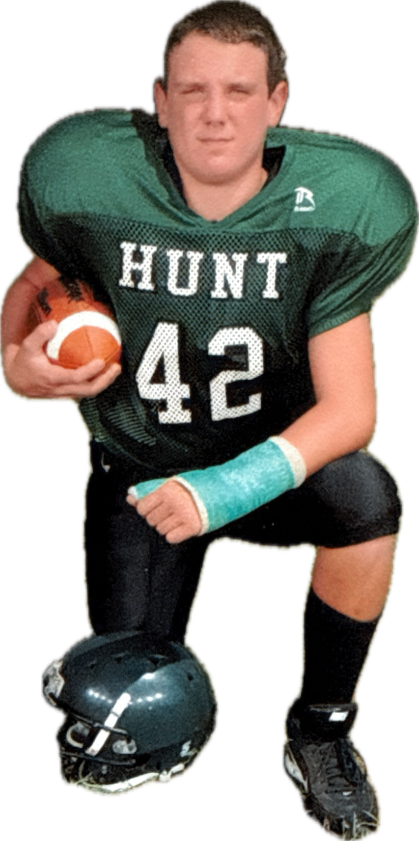 A young male football player in a green and black uniform with the name 'HUNT' and number 42, holding a football, with a football helmet on the ground at his feet, kneeling on one knee with a clenched fist.