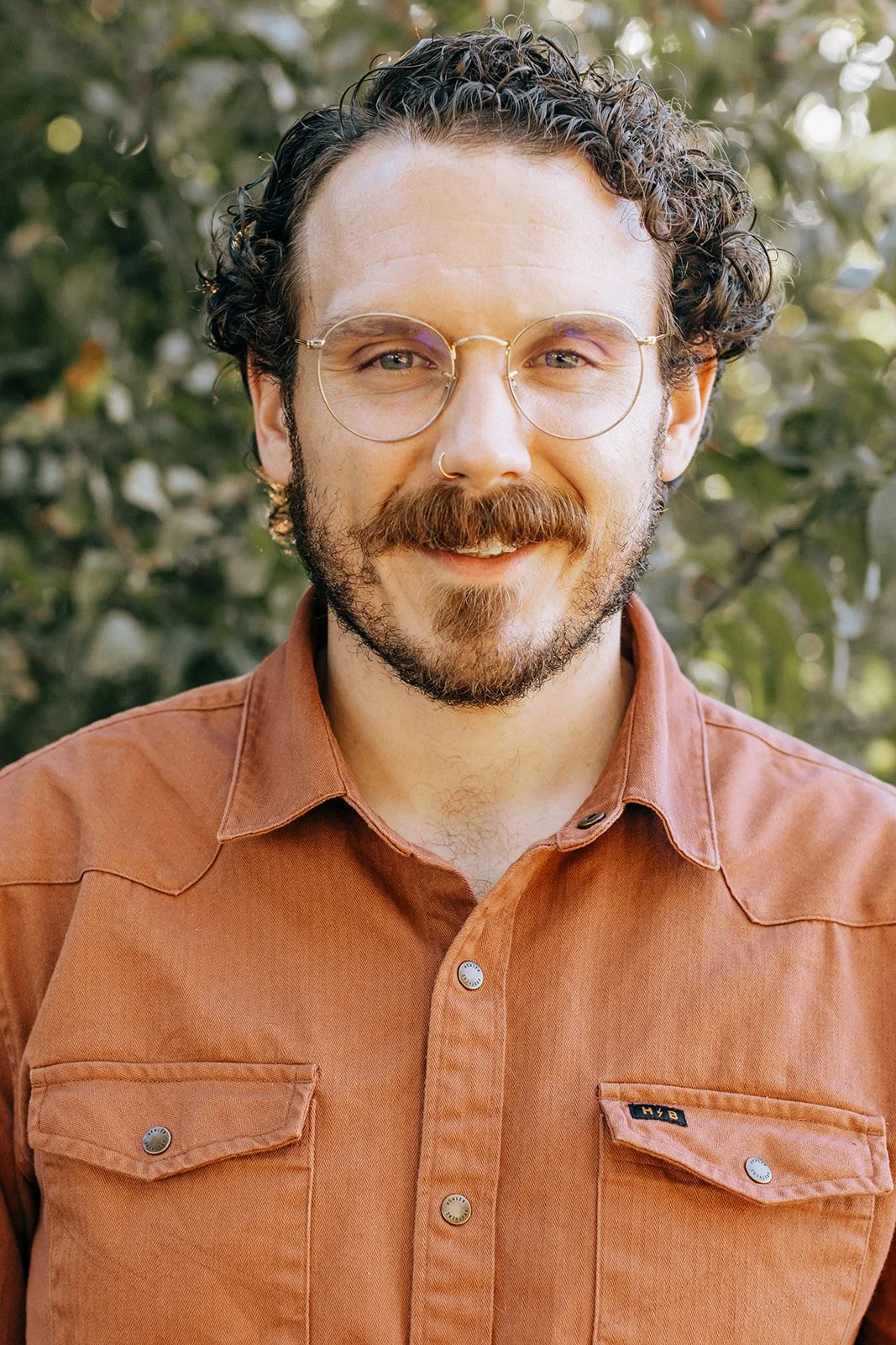 A smiling man with curly hair, glasses, and a beard, wearing a rust-colored button-up shirt, outdoors with greenery in the background.