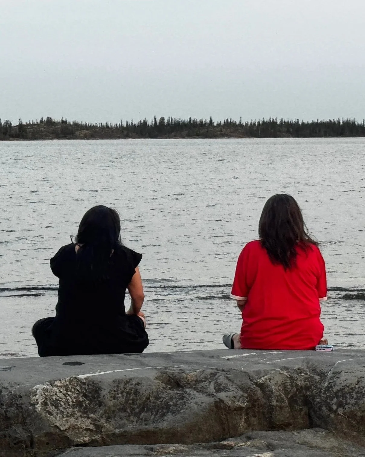 Two women sitting on a stone ledge by a lake, facing the water, with trees across the lake in the distance.