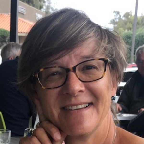 Smiling woman with short blonde hair, wearing glasses with a colorful frame, a beige top with ruffle details, and a gold necklace.