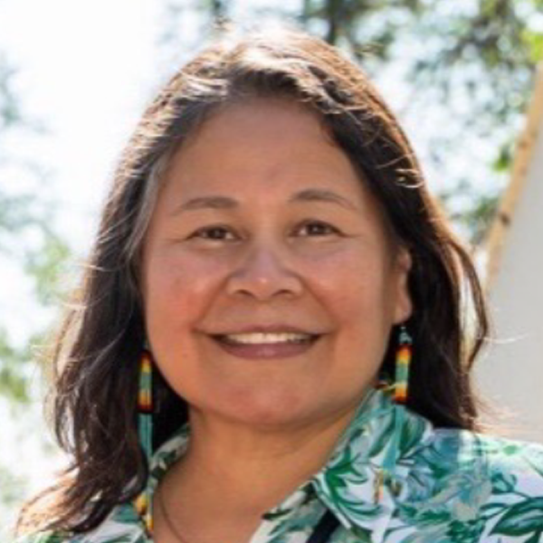 A smiling woman with long dark hair wearing colorful earrings and a floral shirt outdoors, with trees and a structure in the background.