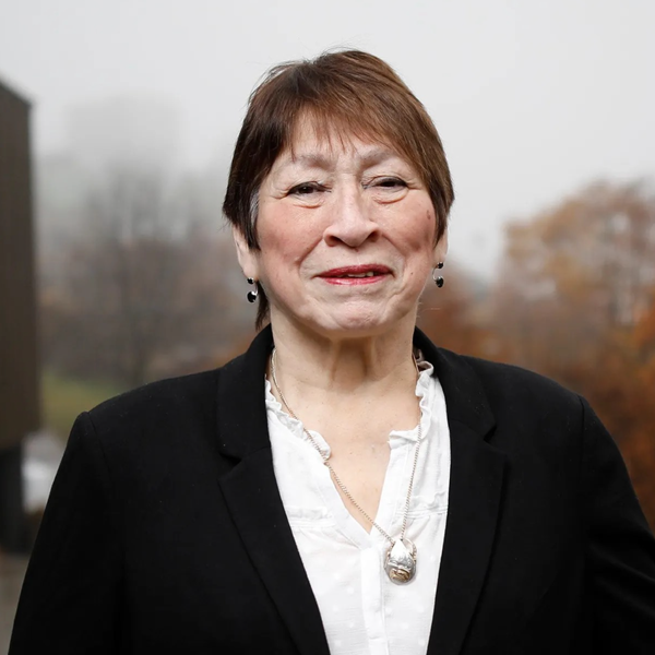 A portrait of an elderly woman with short brown hair, wearing a black blazer over a white blouse with a pendant necklace, standing outdoors with a blurred background of autumn trees.