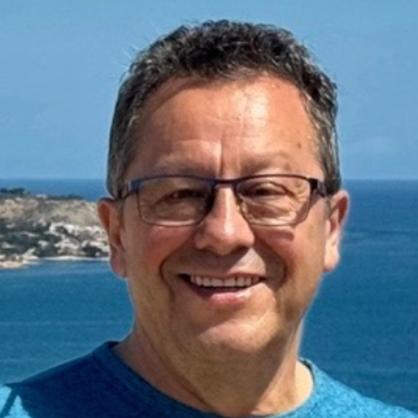 A smiling man with glasses standing outdoors near the coast, with water and a coastline visible in the background.