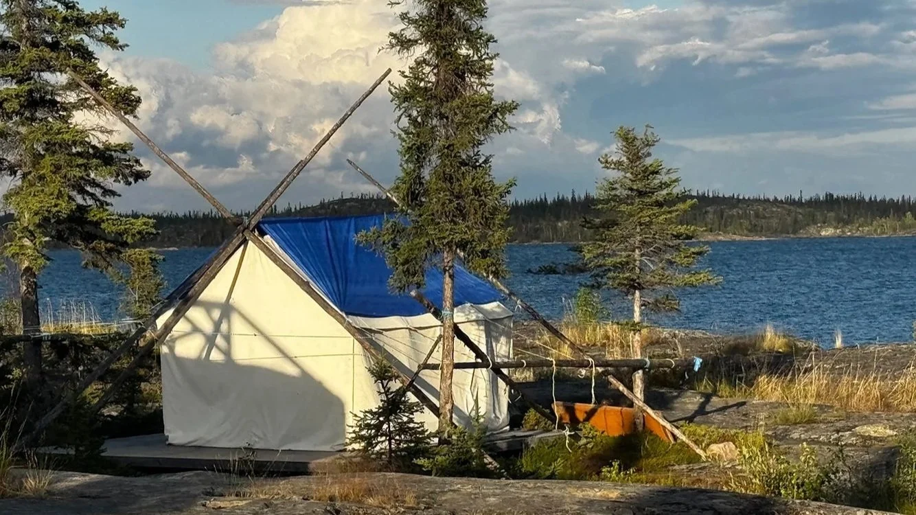 A small canvas wall tent with a blue tarp on the roof set up on rocky ground next to a lake, with trees surrounding it and a cloudy sky overhead.