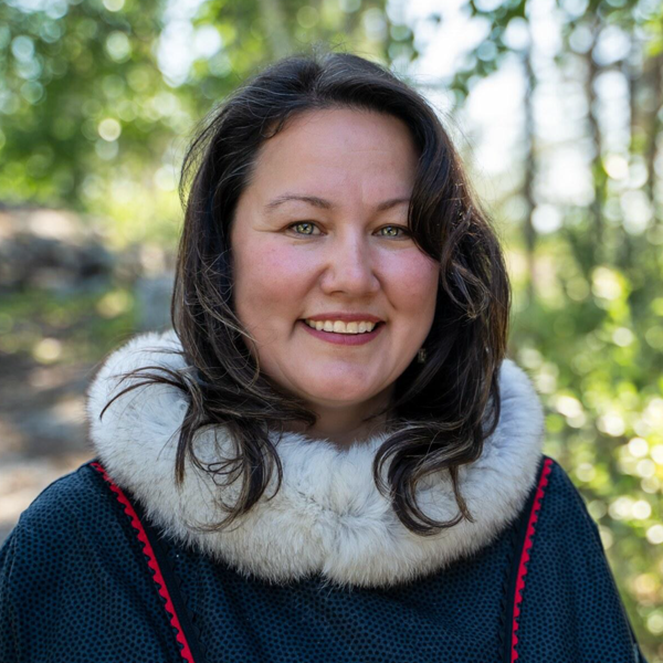 Woman with dark brown hair smiling outdoors in a wooded area, wearing a dark blue top with a white fuzzy collar