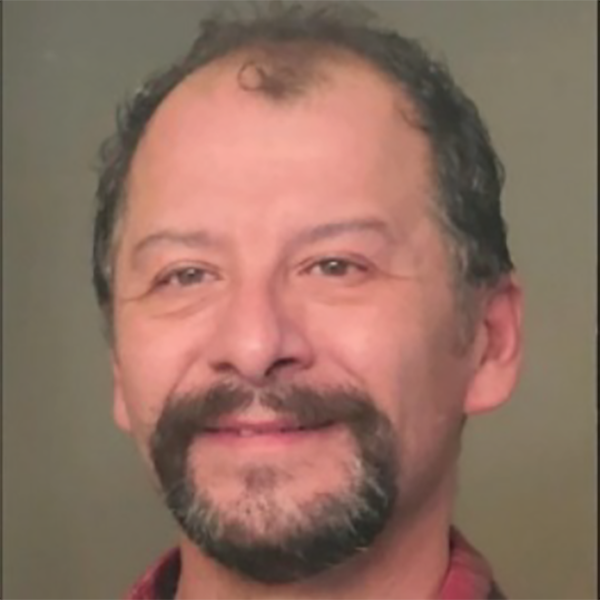 A middle-aged man with short curly hair, a beard, and a mustache, smiling at the camera with a neutral background.