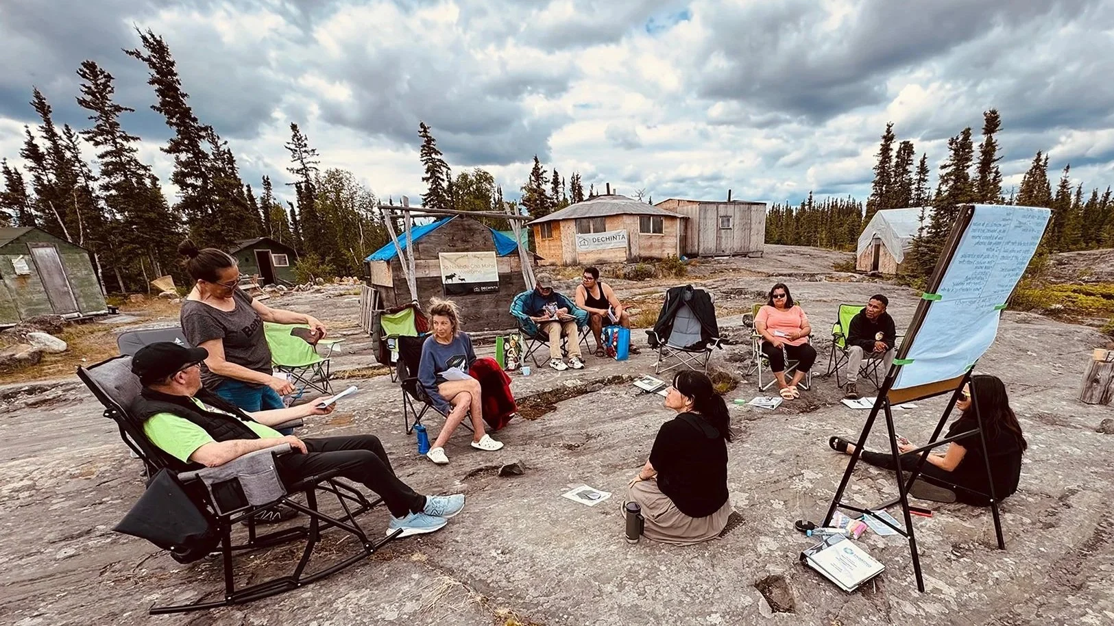 A group of people sitting in a circle outdoors on rocky ground, participating in a discussion or workshop, with some taking notes and a whiteboard nearby.