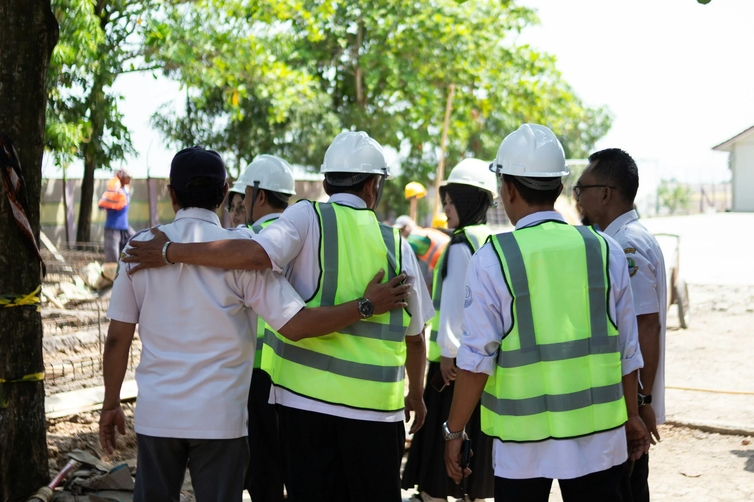 Group of construction professionals wearing safety vests and helmets gathered outdoors, discussing site plans.