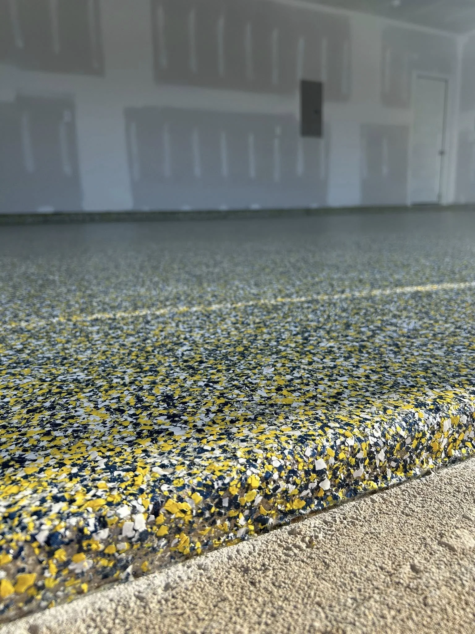 Close-up view of a textured, multicolored epoxy garage floor with black, yellow, and white speckles, with a garage door and white wall in the background.