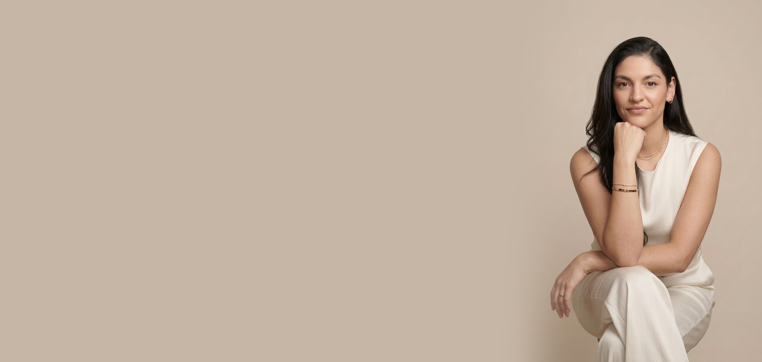 Portret van een jonge vrouw met lang zwart haar, zittend en rustend haar kin op haar hand, gekleed in een witte outfit, voor een beige achtergrond.