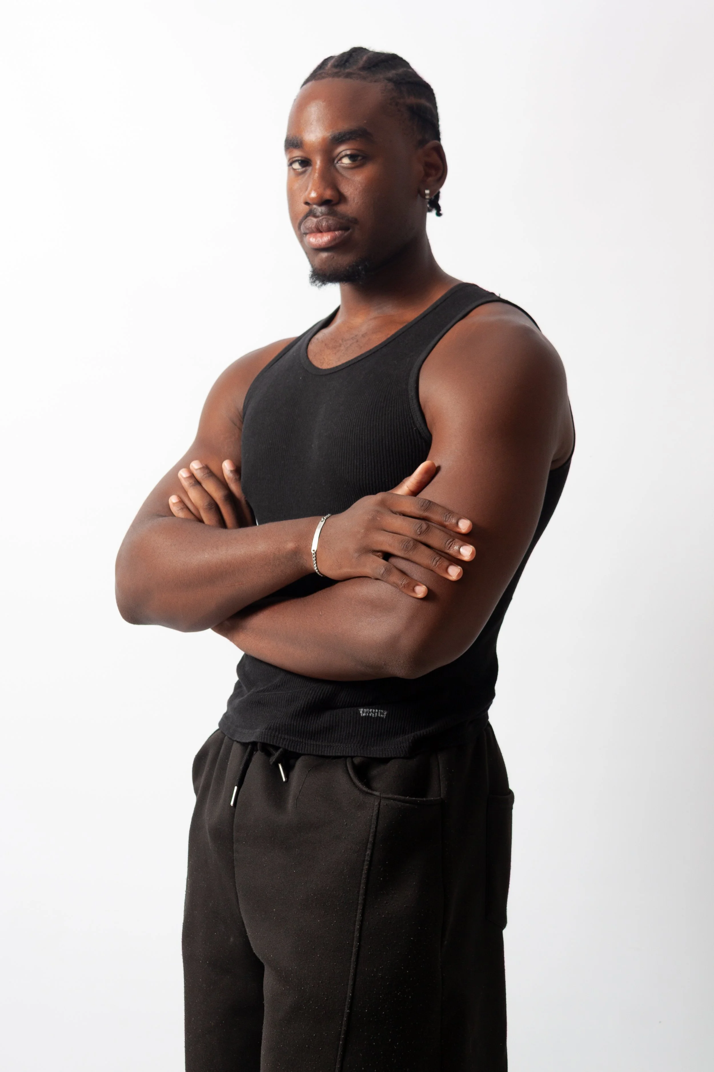 A young man with braided hair and earrings, wearing a black tank top and black sweatpants, standing with crossed arms against a white background.
