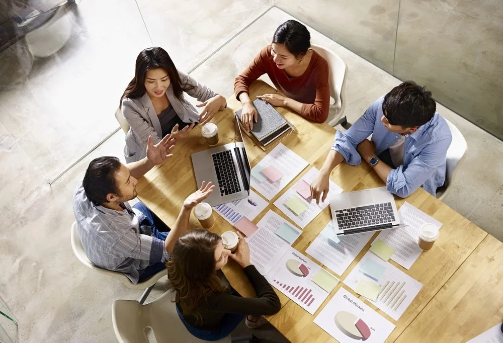 A group of six diverse people having a discussion around a wooden table in a modern office. They are surrounded by laptops, documents, pie charts, and sticky notes. Some are holding coffee cups, and one is gesturing while speaking.
