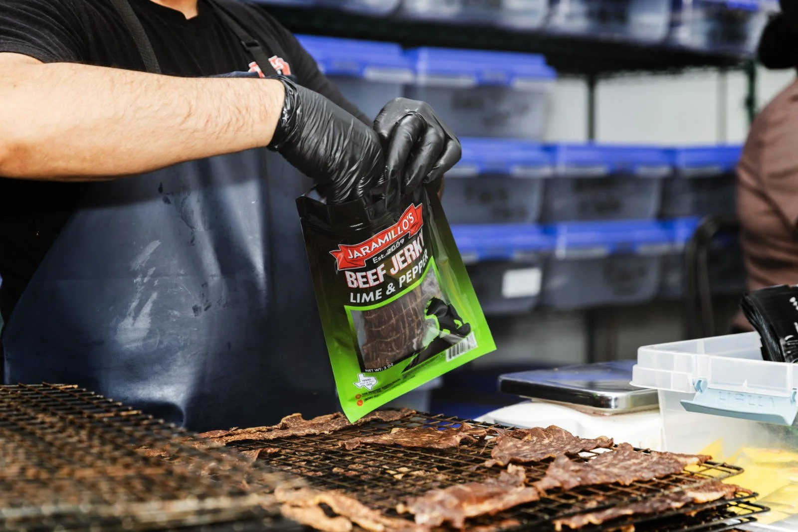 Person wearing black gloves and apron places bag of beef jerky on grill with cooked meat strips. Shelves with plastic containers are in the background.
