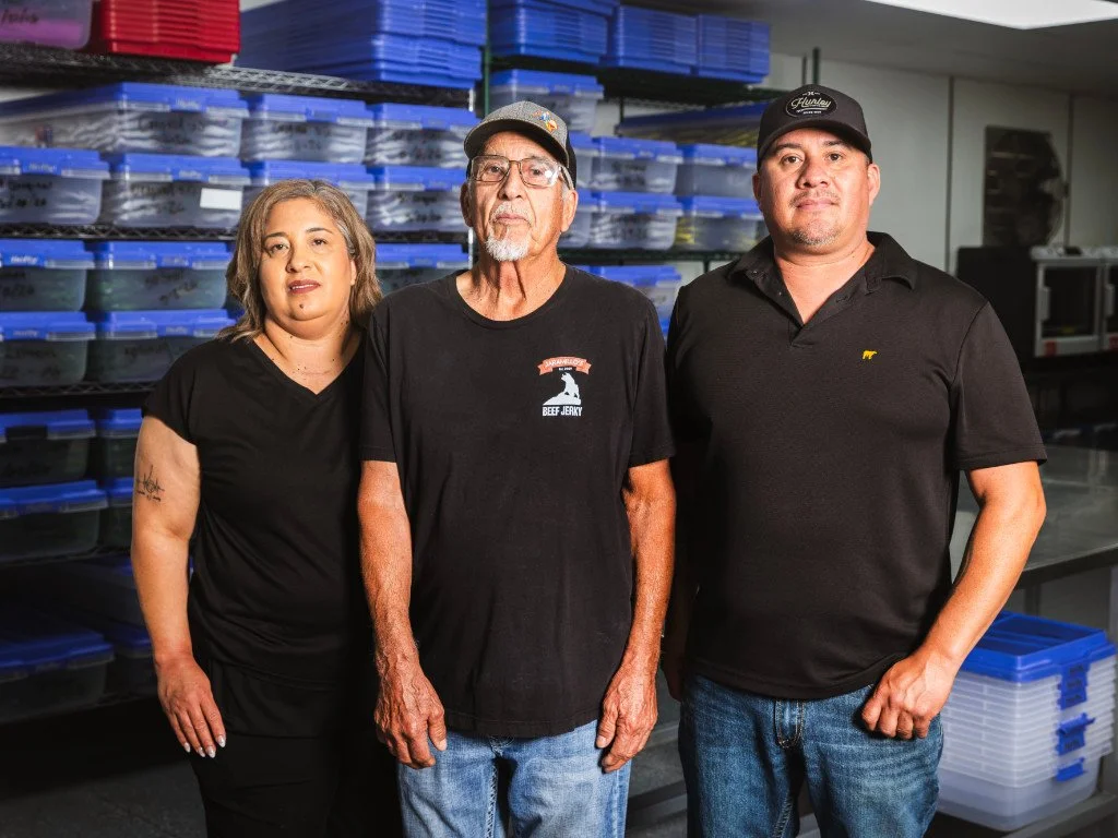 Three people standing in front of shelves filled with plastic containers in a commercial kitchen or storage area.