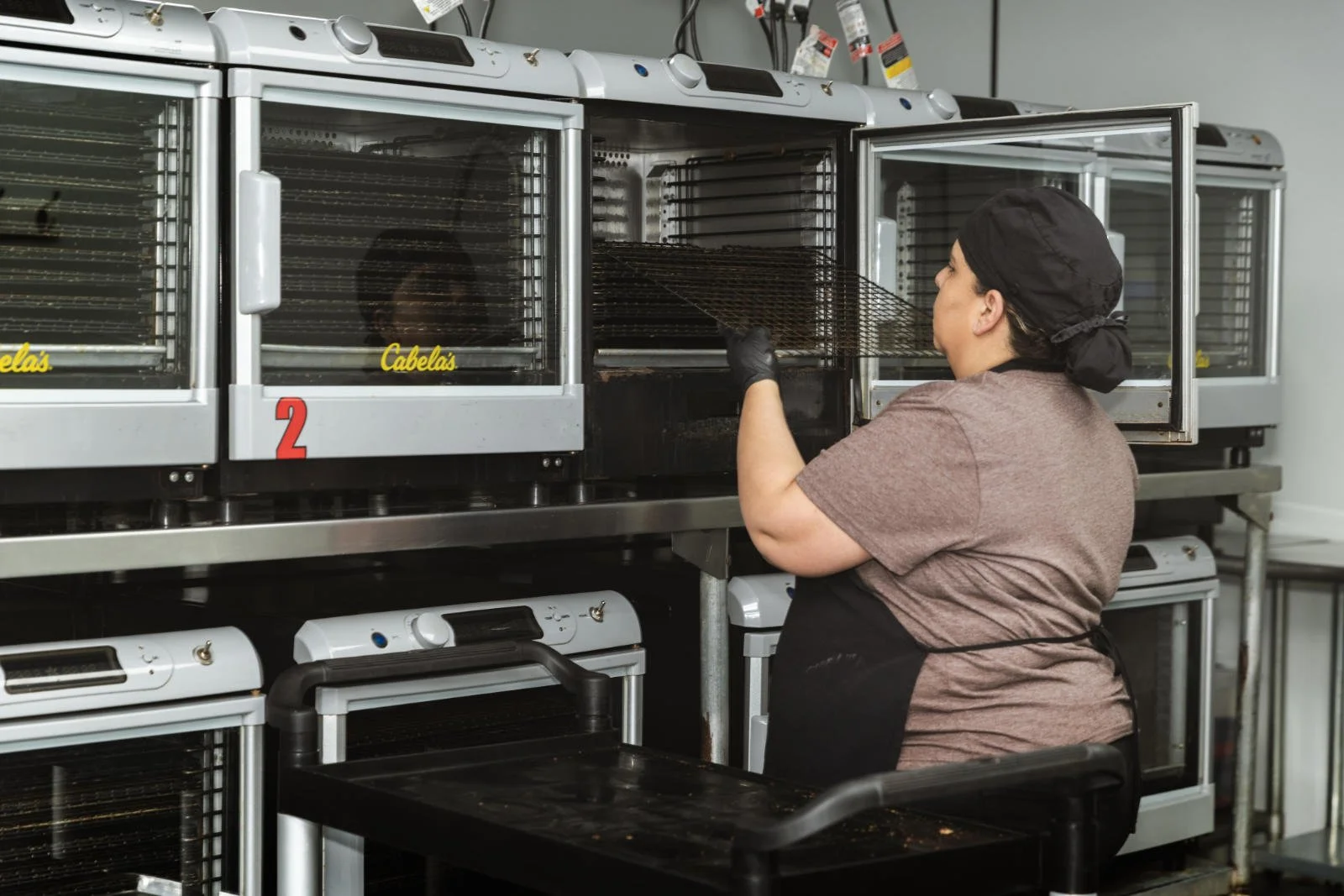 A woman wearing a black hair covering, brown shirt, and black apron cleaning the inside of a commercial-grade commercial smoker or oven with a wire rack.