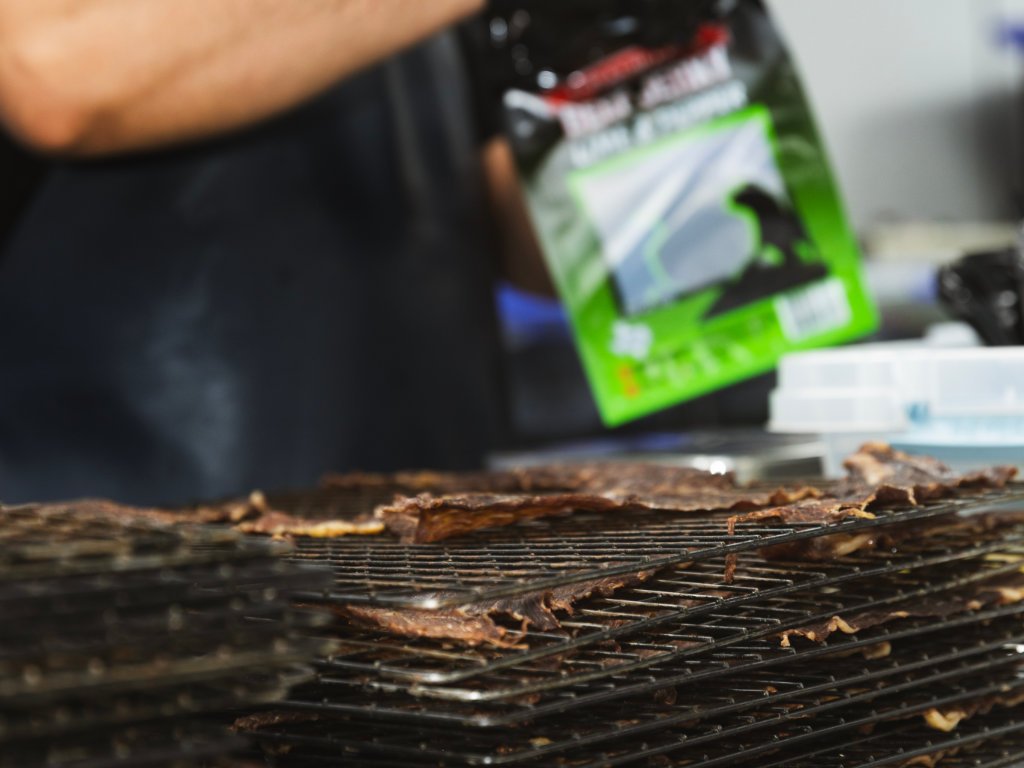 Close-up of bacon slices cooking on a wire rack, with a blurred background including a black package and some white containers.