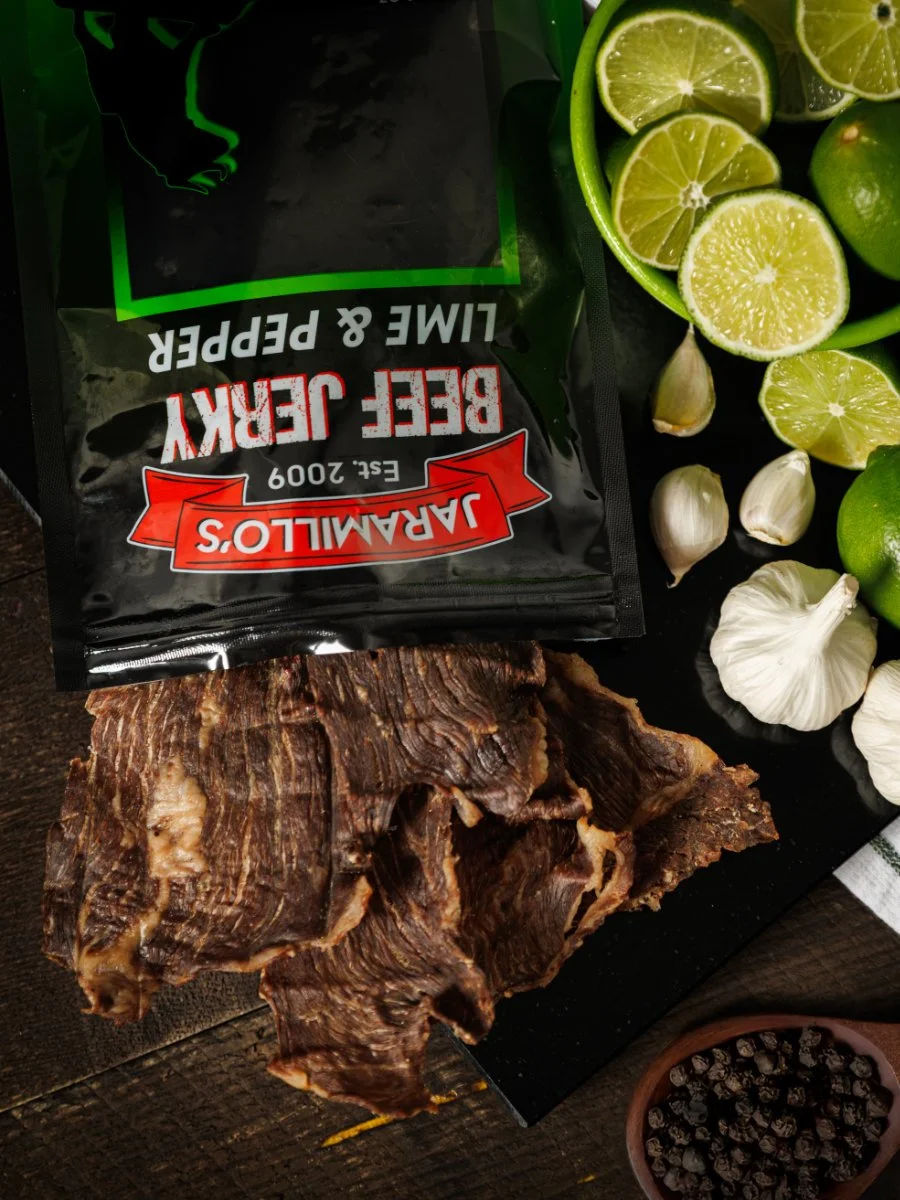 Dried beef jerky, garlic cloves, lime halves, black peppercorns, and a packet of Jaramillo's Bee-Jerky Lime & Pepper beef jerky on a wooden surface.