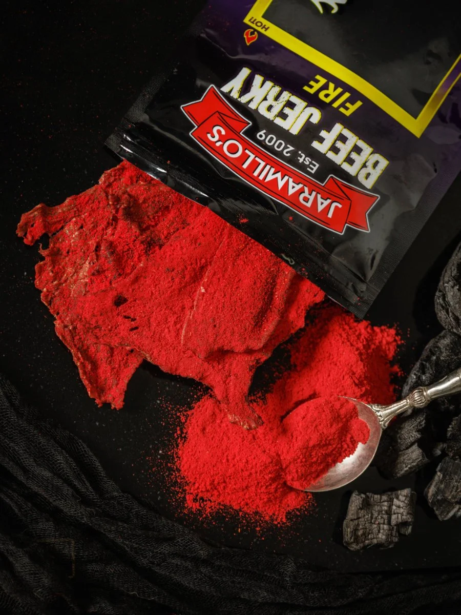 A black surface features a partially opened red and black packet labeled "JARALLIOS BEER JERKY" with the text "EST. 2009" and "FLAVOR." Next to the packet, there is a large piece of red powdered seasoning and a silver spoon filled with the red powder. Charcoal pieces are visible to the right, and black fabric is around the scene.
