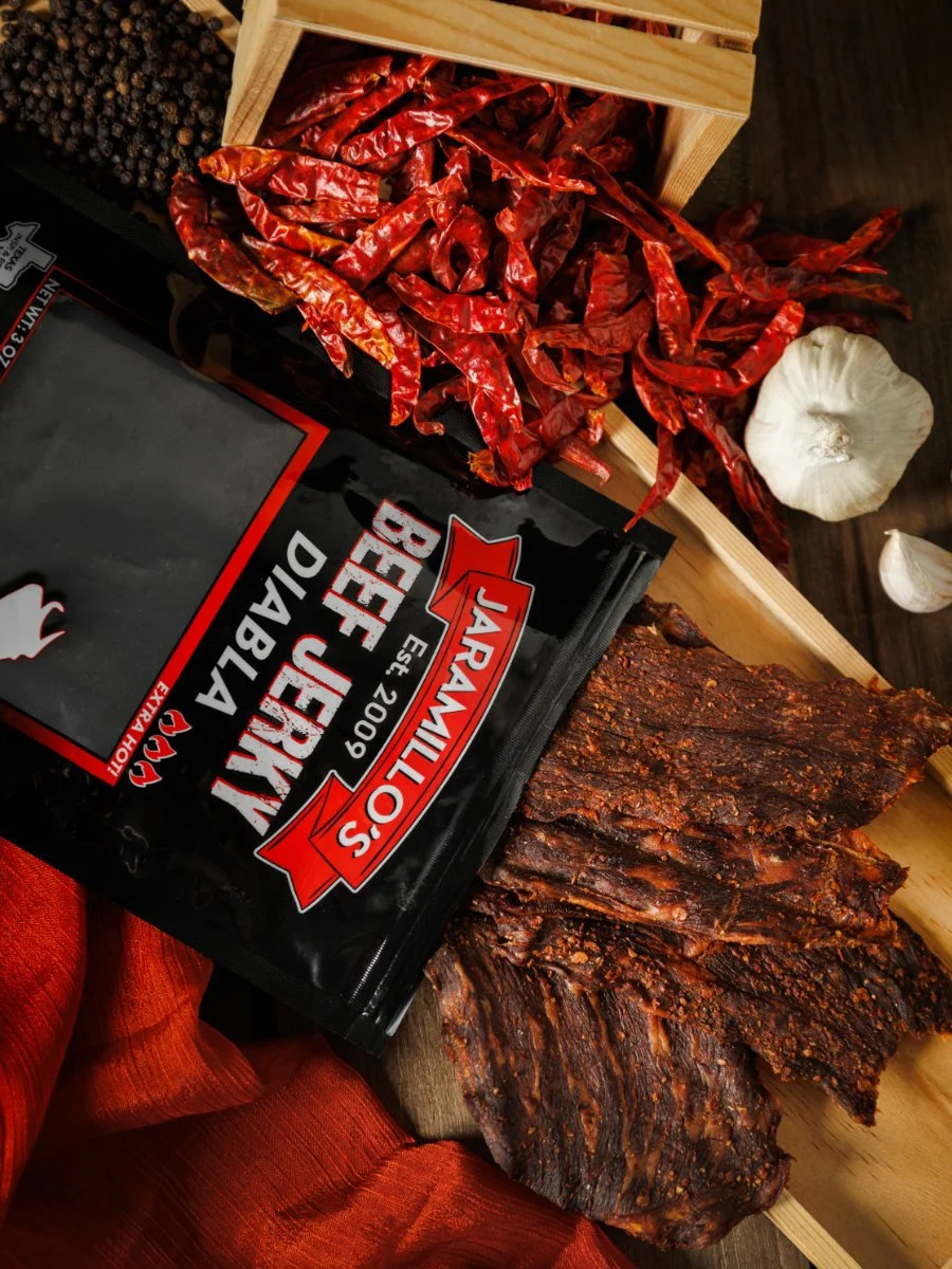 Smoked beef briskets, dried red chili peppers, garlic, black peppercorns, and a package of Jaramillo's beef jerky on a table.