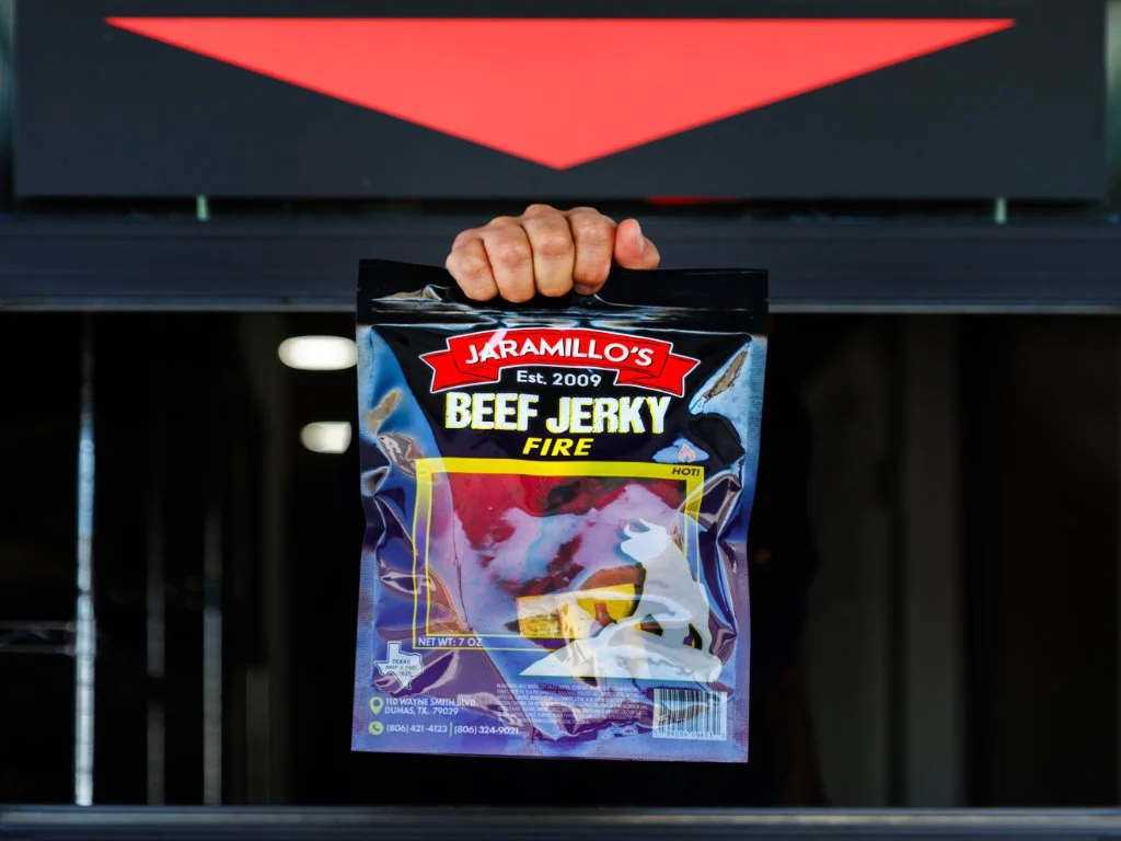 A hand holding a bag of Jaramillo's Beef Jerky Fire flavor, with bright packaging that features the brand name, flavor, and a warning label for hot spice.